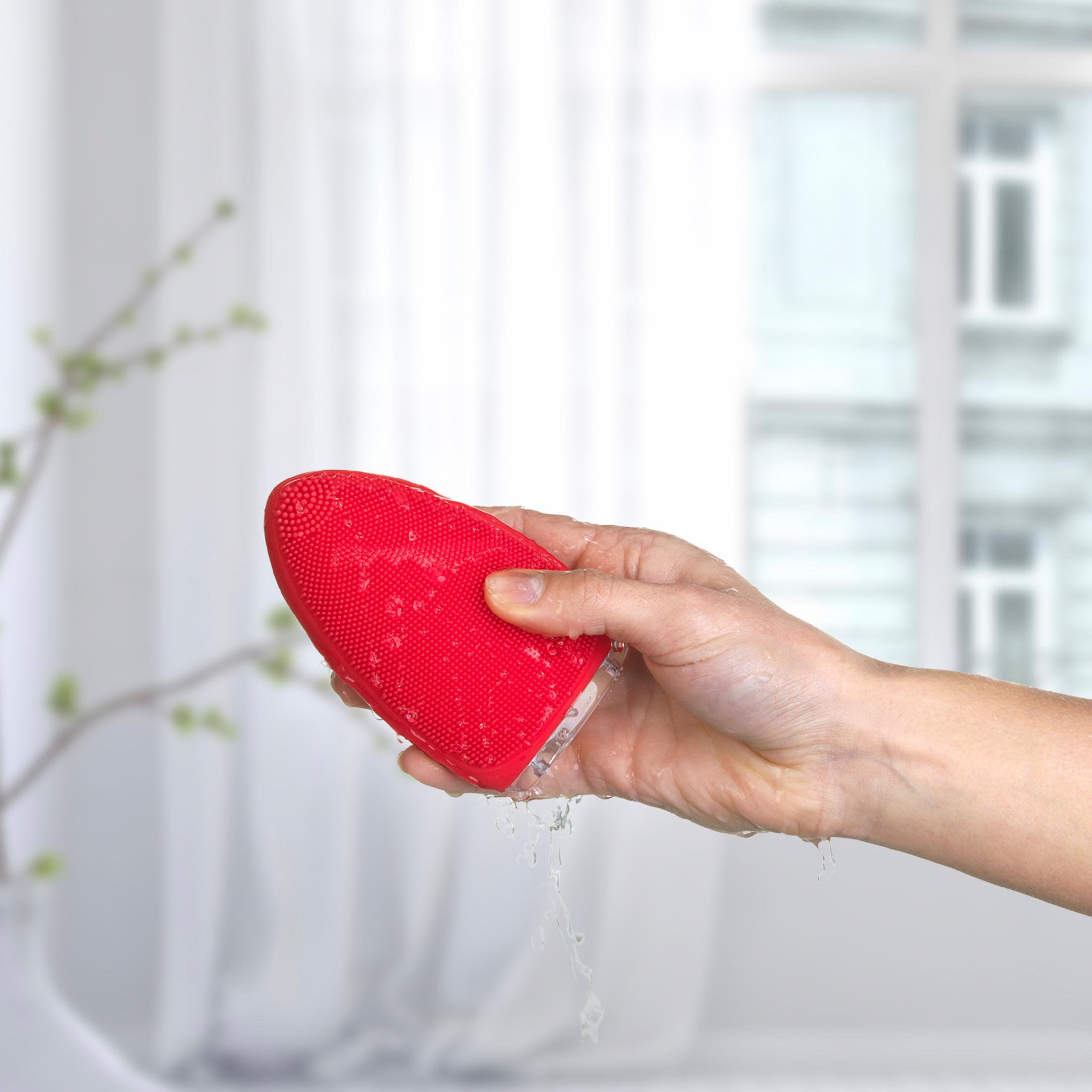 Una mano sostiene un cepillo de limpieza facial de silicona roja con agua goteando.