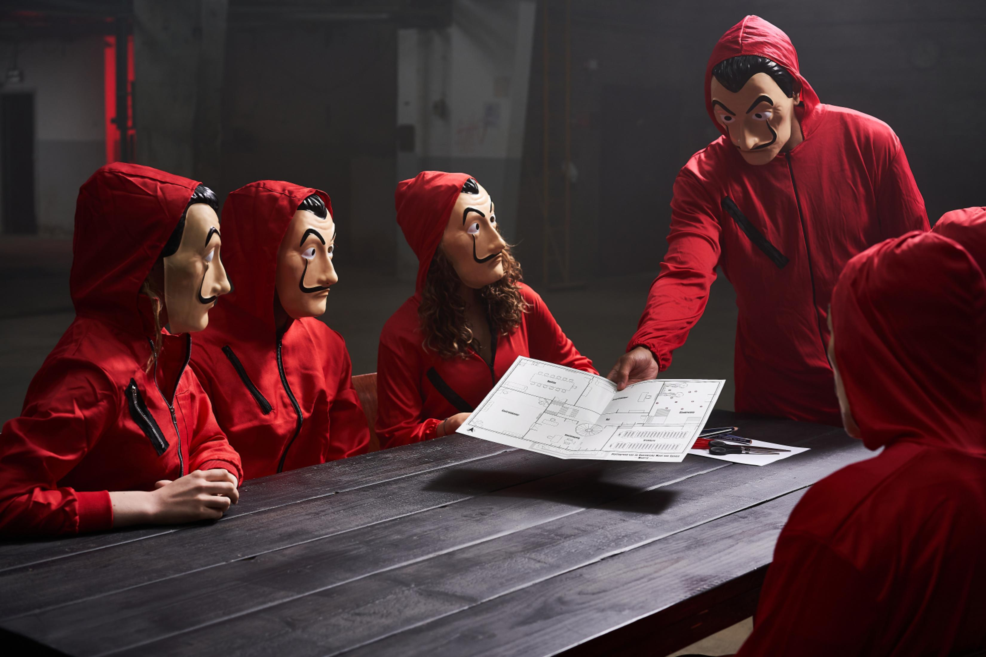 Personen in roten Overalls und Dali-Masken sitzen um einen Tisch und betrachten einen Bauplan.