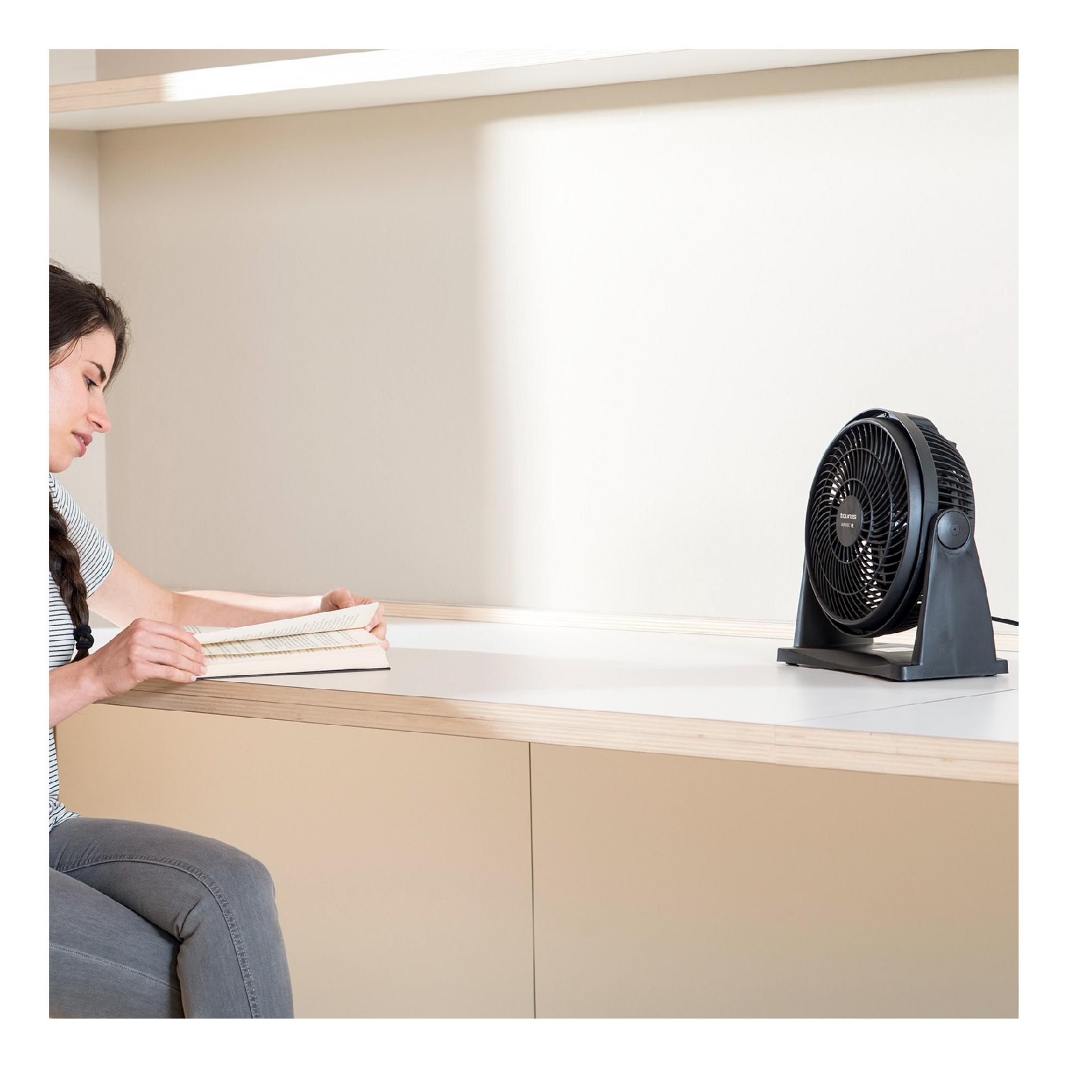 Frau liest Buch, ein schwarzer Ventilator steht auf dem Tisch.