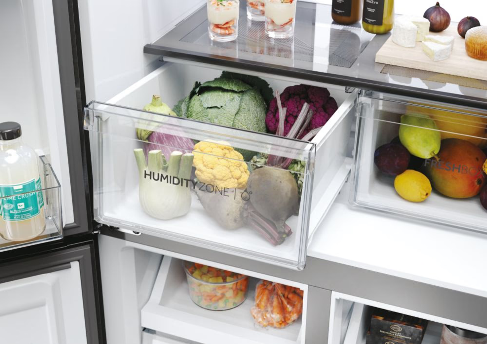 Ein Kühlschrank mit Gemüse und Obst in Schubladen. Unten ist eine Flasche Limonade.