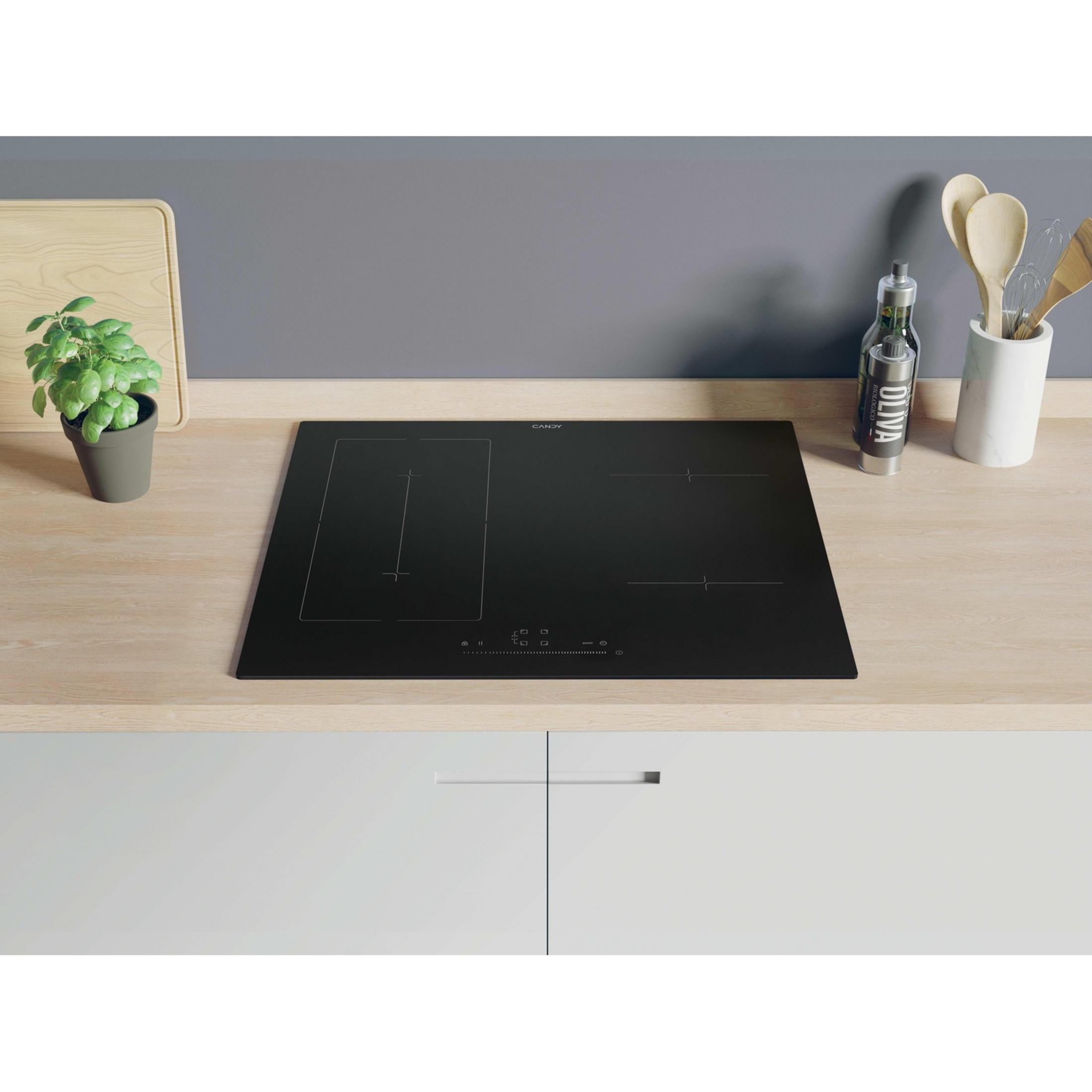 A black induction hob on a wooden surface with kitchen utensils and a plant.