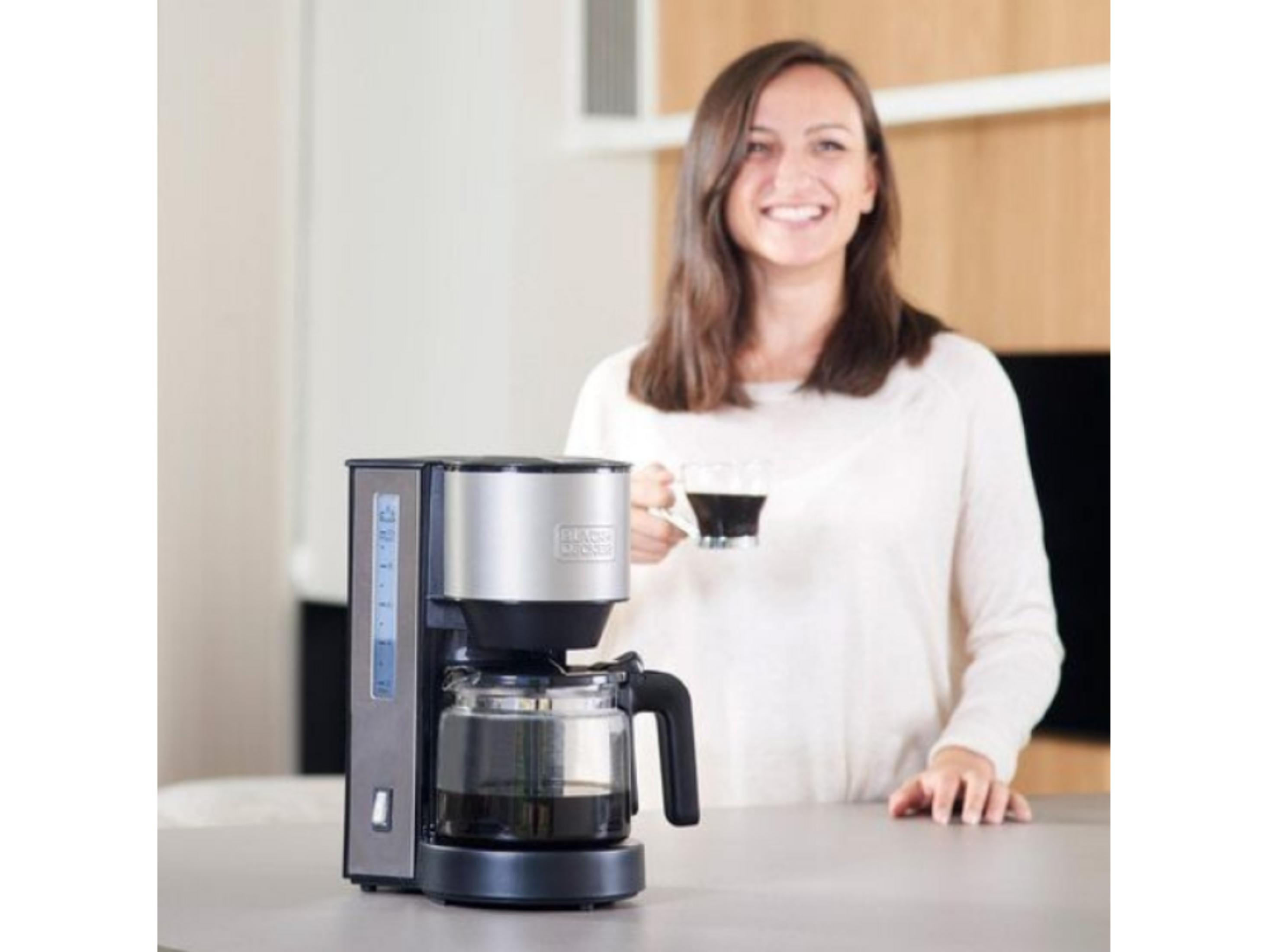 Una mujer sostiene una taza de café junto a una cafetera.