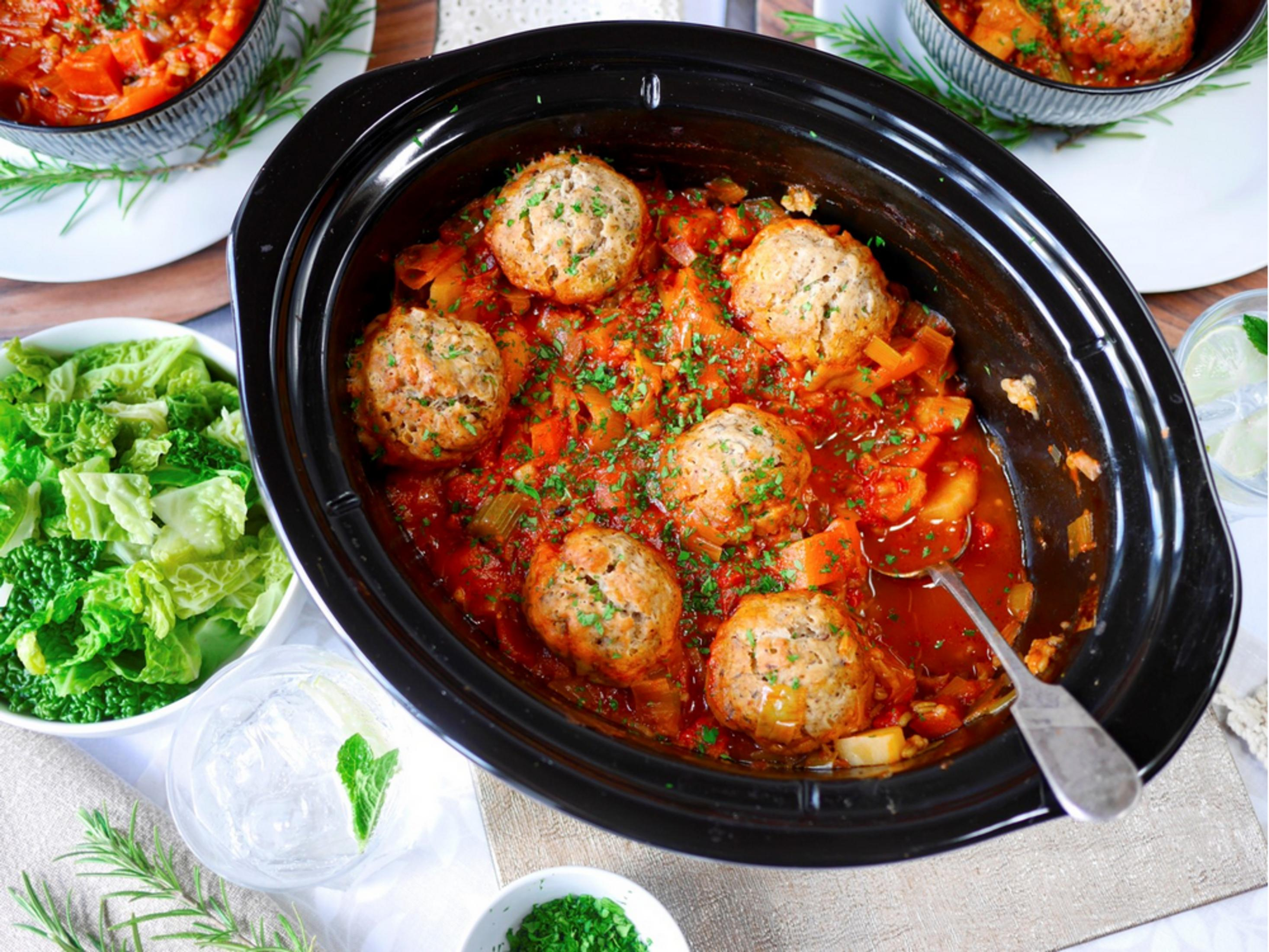 Ein schwarzer Schmortopf mit Klößen in Tomatensoße, daneben Salat und Getränk.