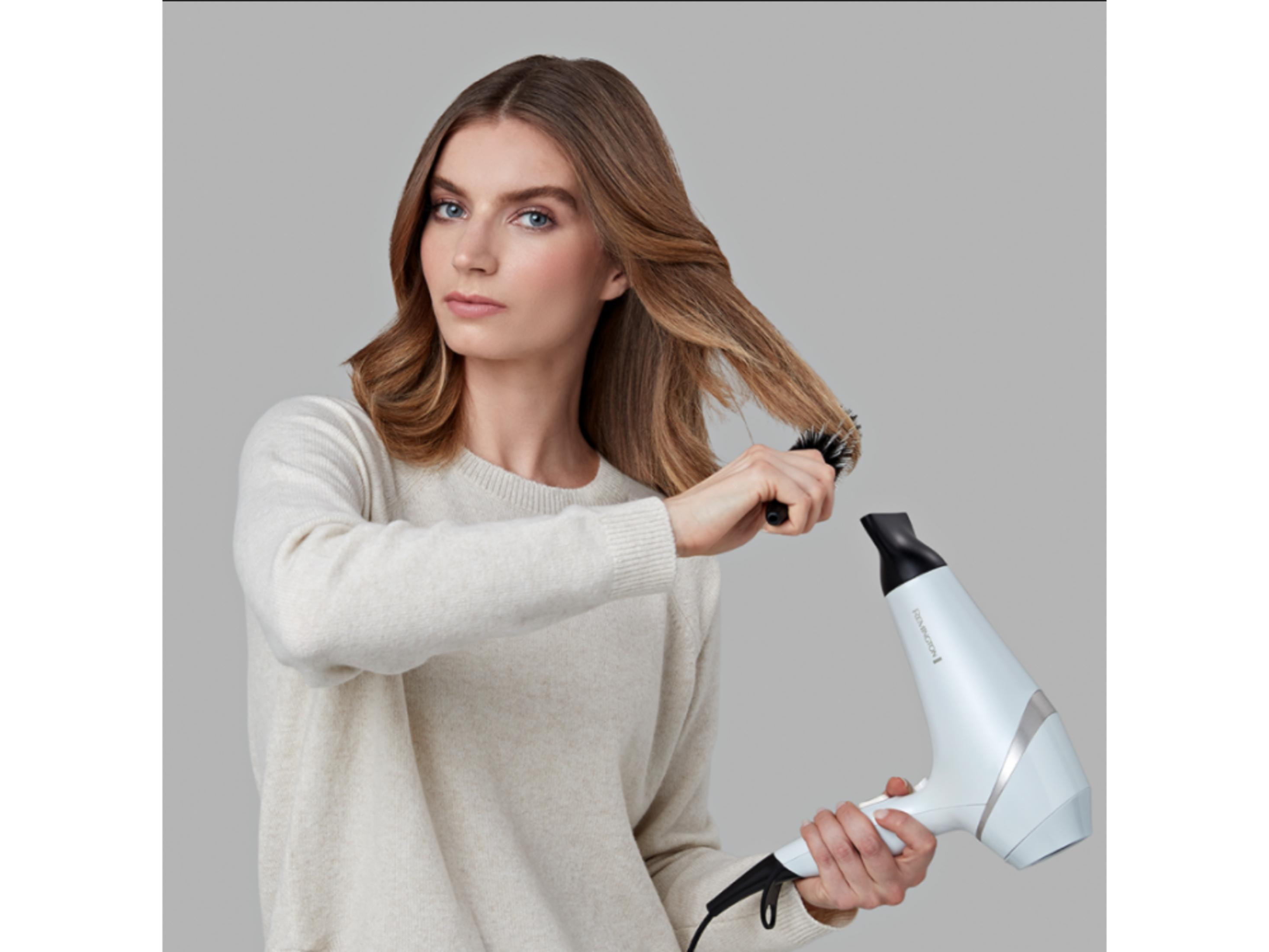Une femme sèche ses cheveux bruns avec un sèche-cheveux Remington blanc et une brosse.