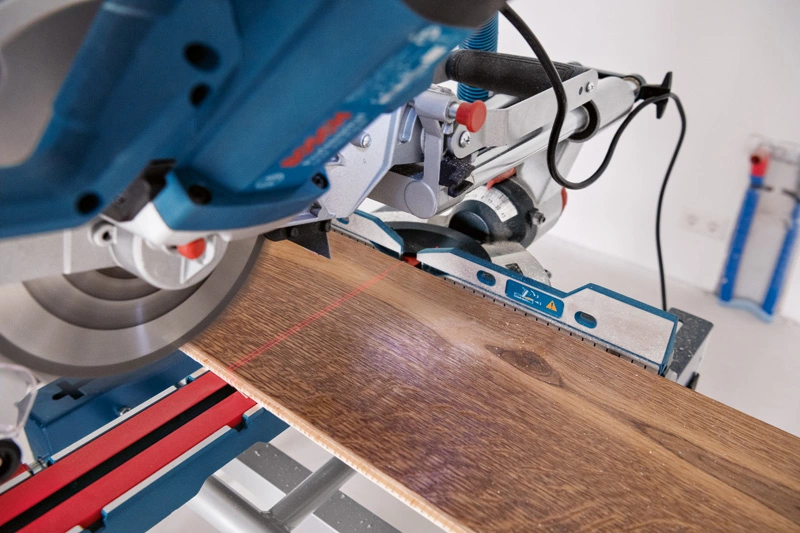 Ein blau-silberner Gehrungssäge schneidet ein Holzbrett mit roter Laserführung.