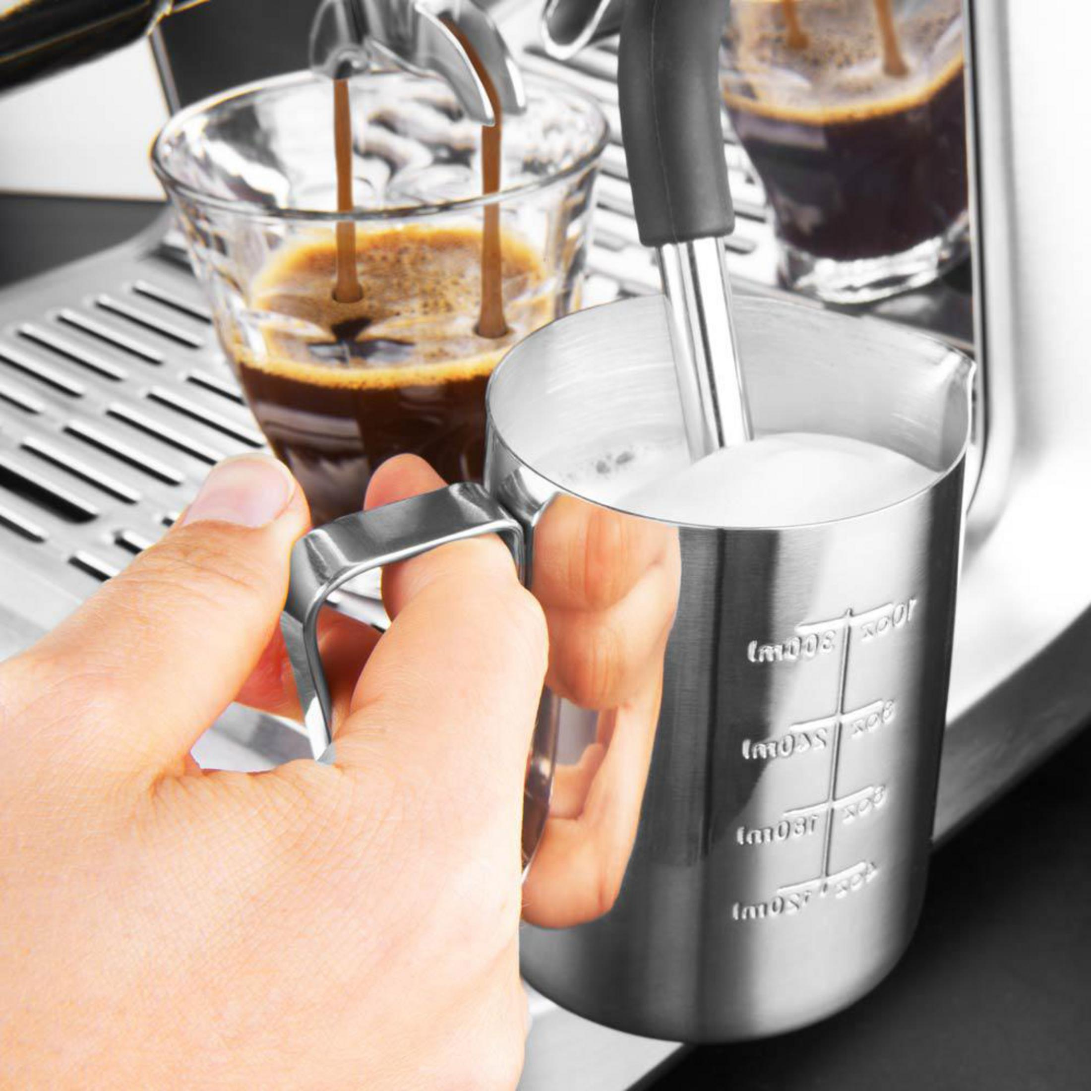Una mano tiene una caraffa di metallo con latte montato accanto a un espresso che cola in un bicchiere.