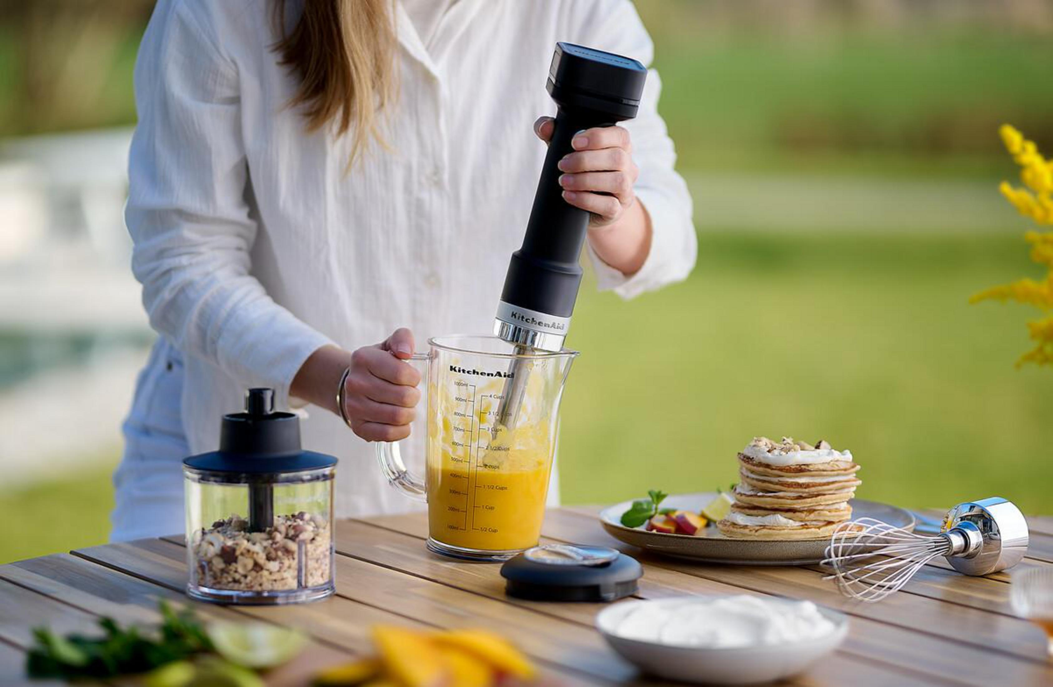 Person benutzt schwarzen Stabmixer in Krug mit orangefarbener Flüssigkeit. Waffeln, Schüssel Sahne auf Tisch.