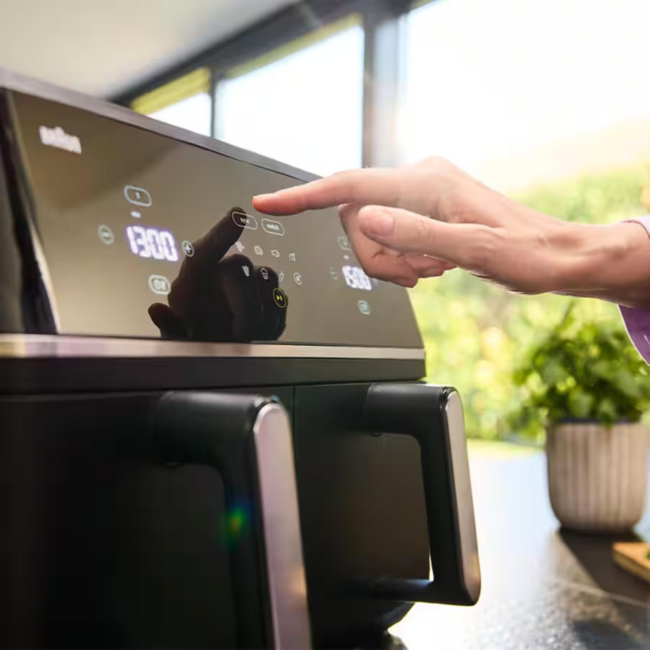Un dito preme un display digitale su un elettrodomestico nero. Vegetazione visibile all'esterno.
