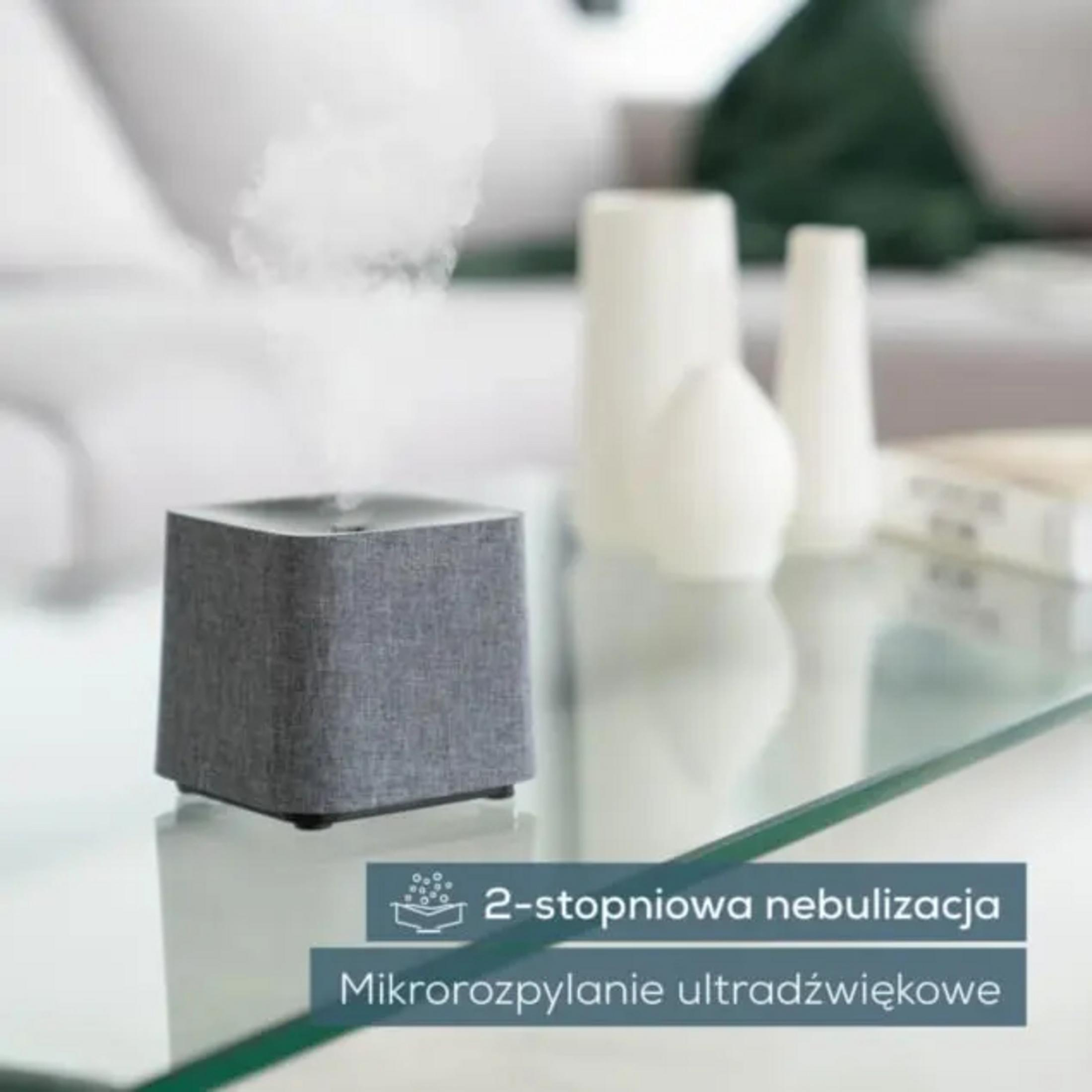 A gray diffuser emits mist, on a glass table with decor objects in the background.