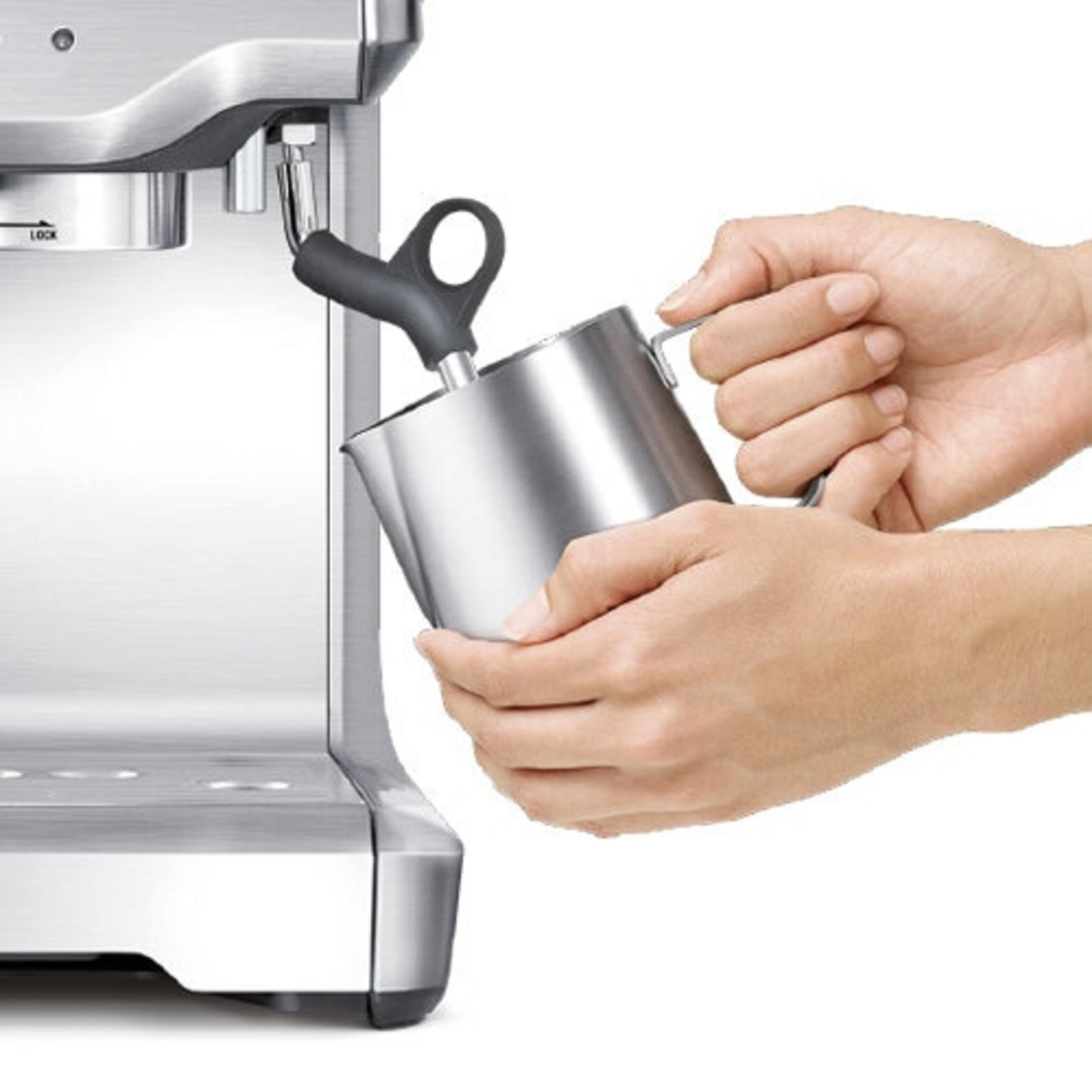Eine Hand hält einen Milchaufschäumerkrug neben einer Kaffeemaschine.