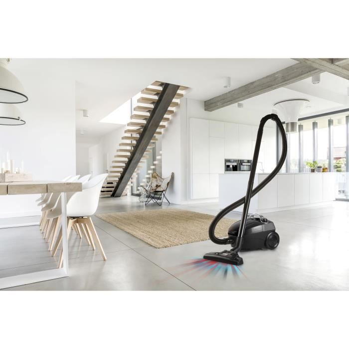 Una aspiradora negra está en un suelo de hormigón en una sala de estar moderna con escaleras.