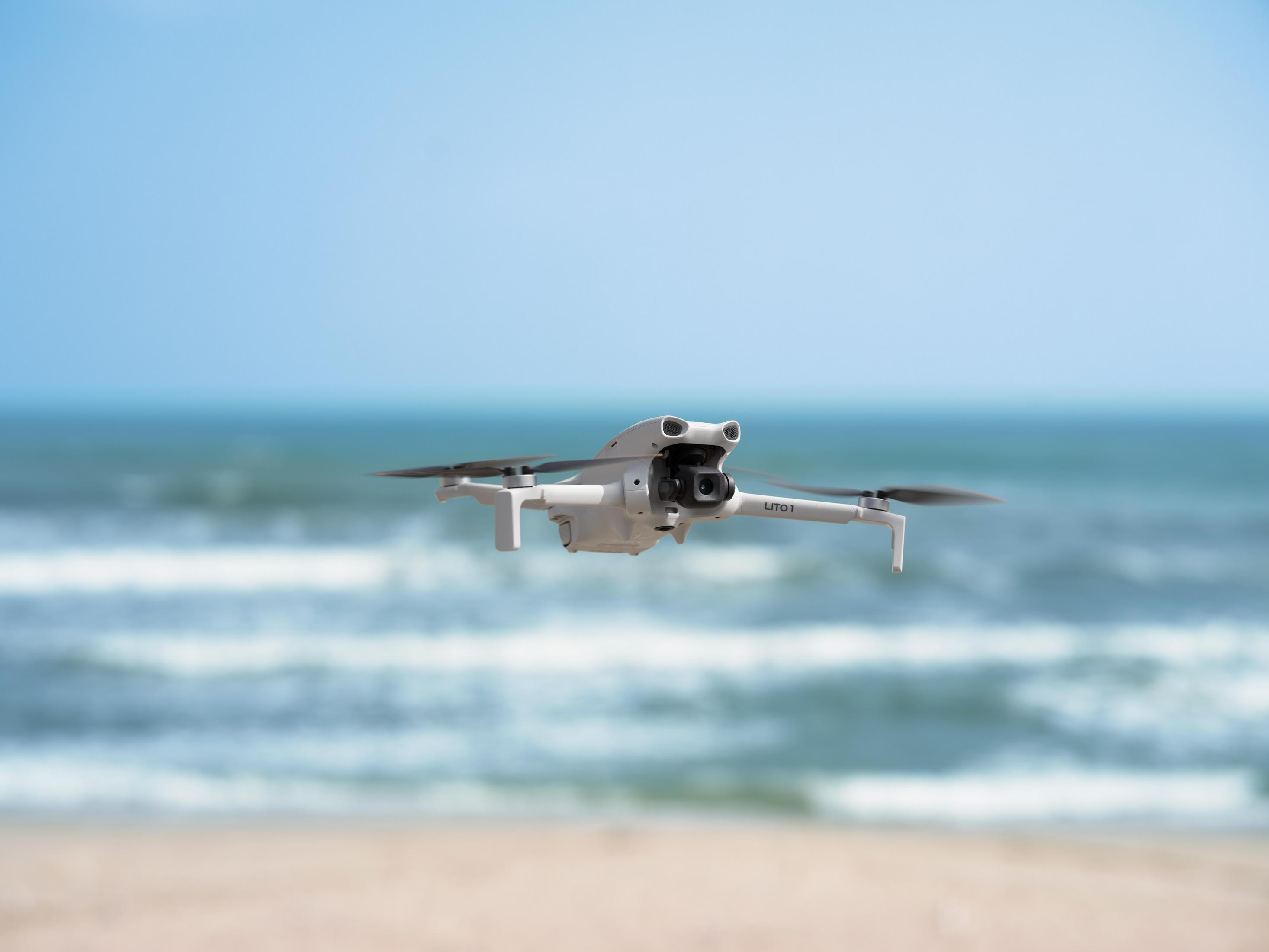 Un dron blanco vuela sobre una playa con un mar azul y olas al fondo.