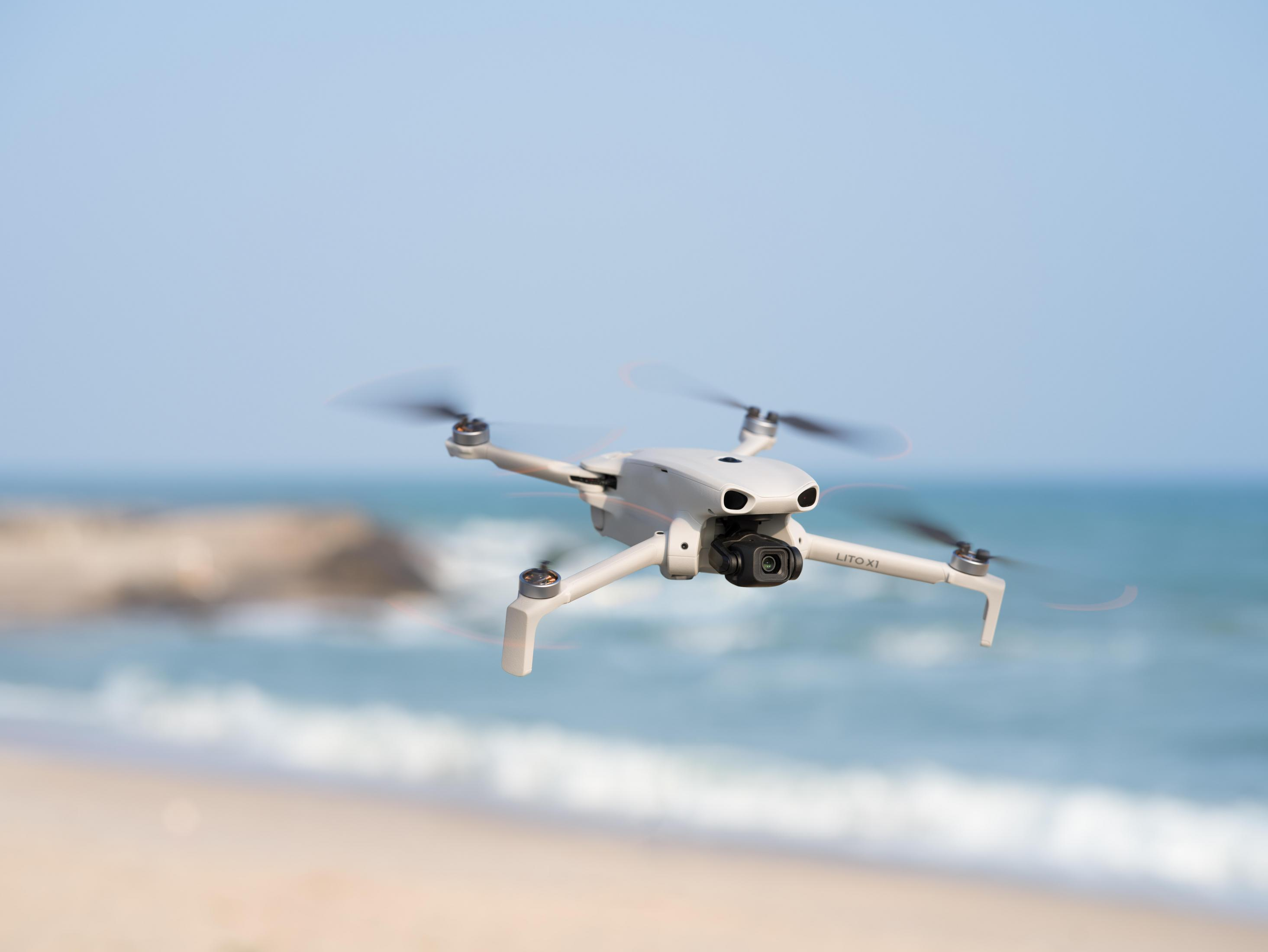 Un dron blanco vuela sobre una playa con agua azul y olas.