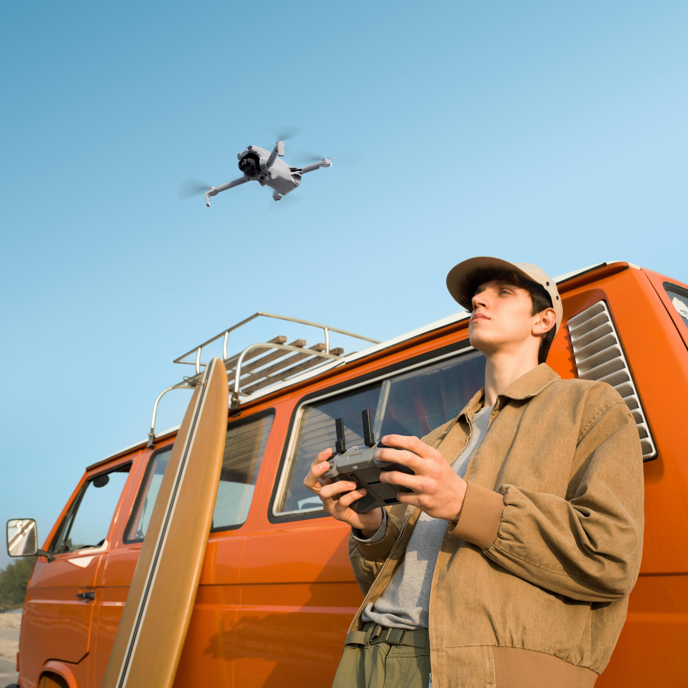 Un hombre pilota un dron frente a una furgoneta naranja con tabla de surf. Cielo despejado.