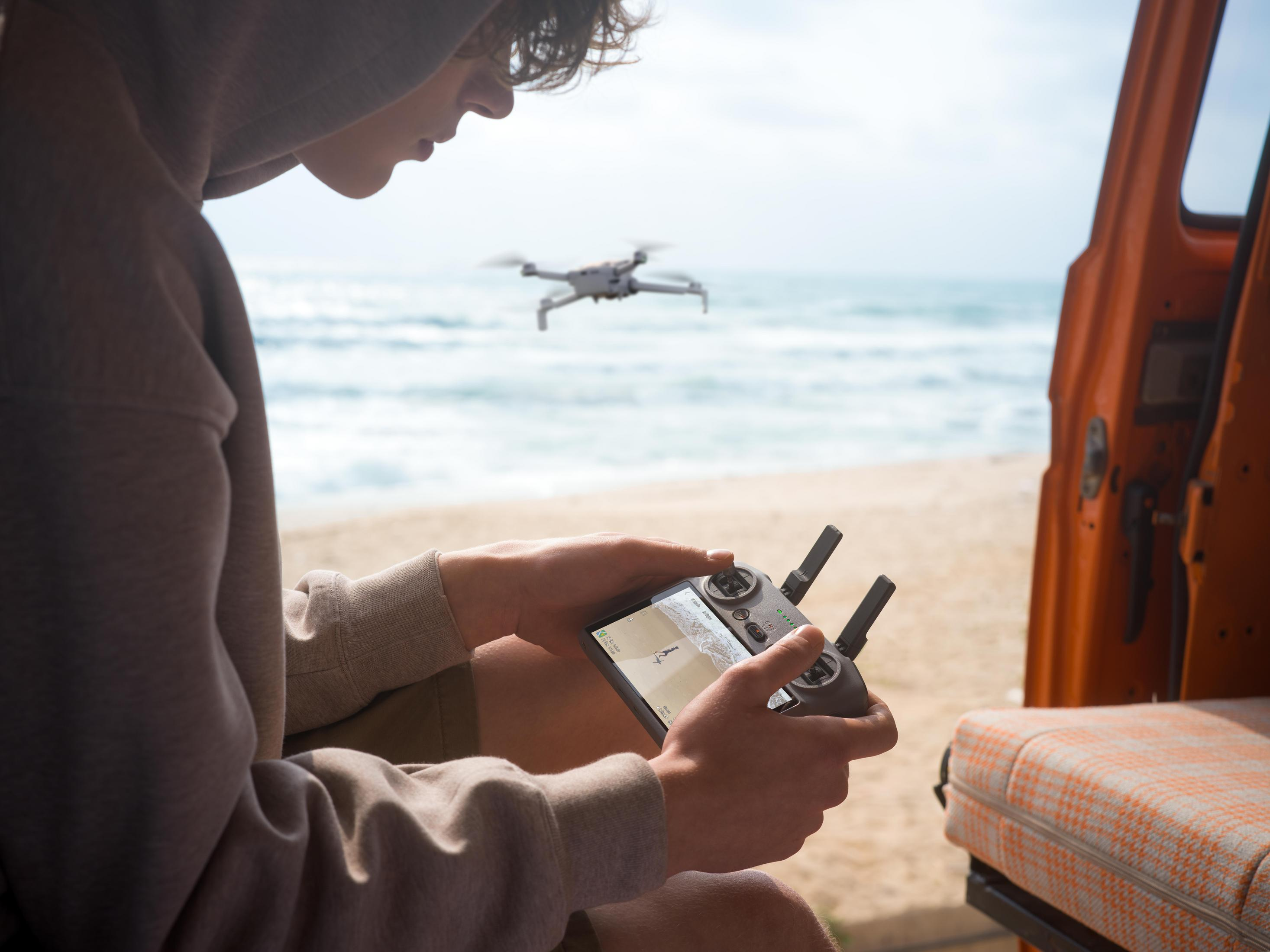 Persona controla dron en playa con mando, mar al fondo.
