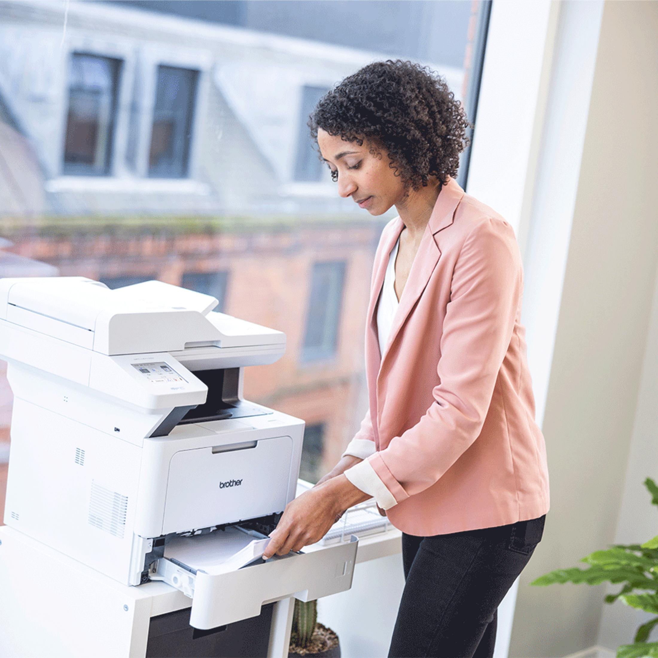 Une femme en blazer rose charge du papier dans une imprimante blanche.