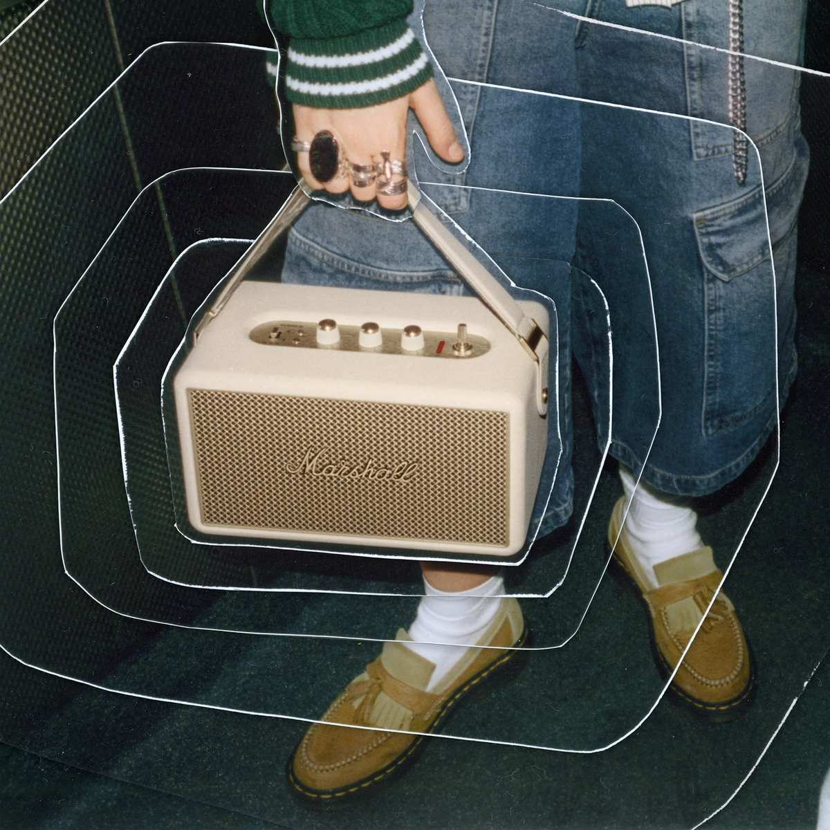 Una persona tiene uno speaker Marshall color crema. Indossa jeans blu, un maglione verde e mocassini marroni.