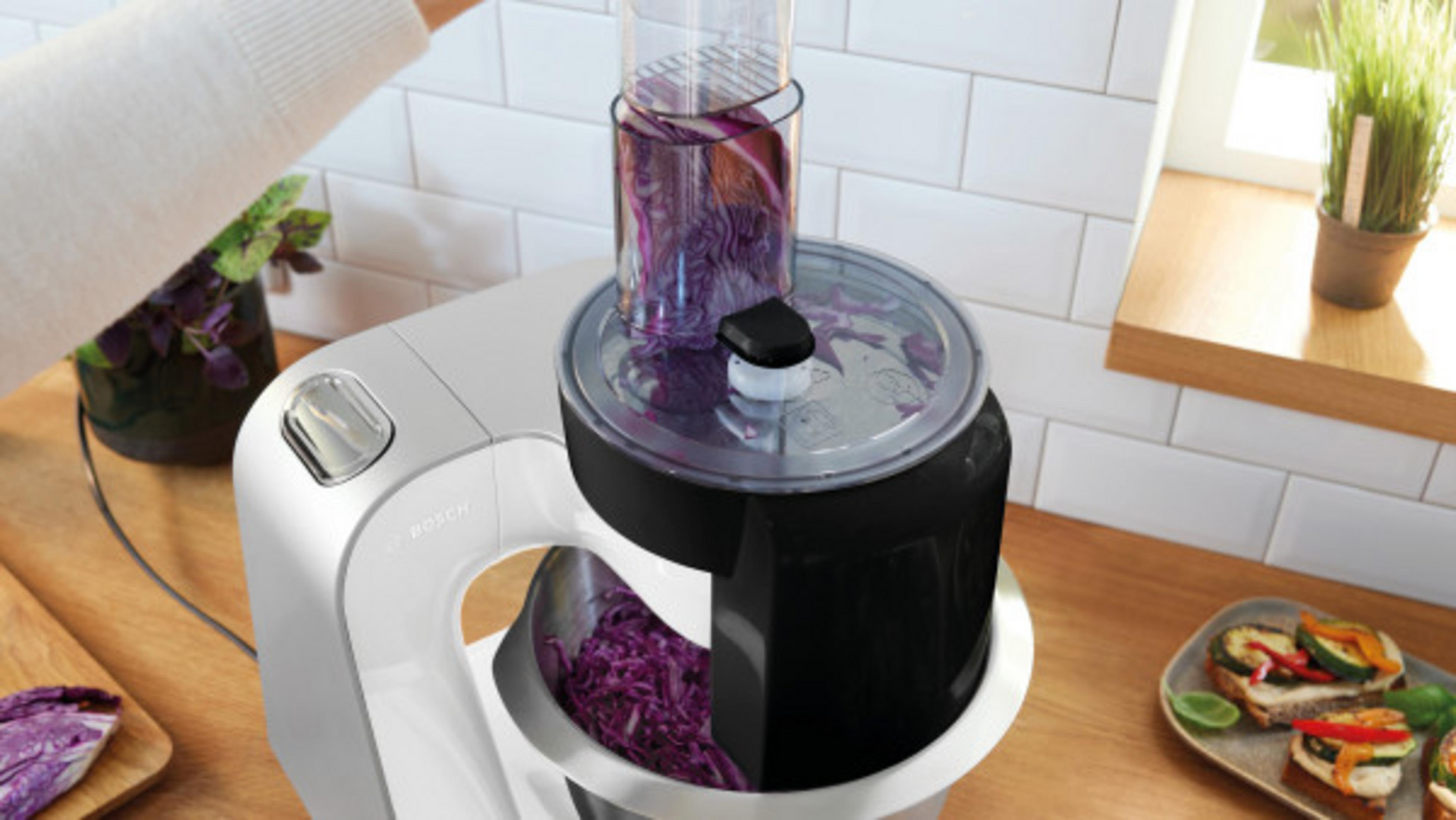 Un robot da cucina lavora il cavolo rosso, con toast e verdure nelle vicinanze.
