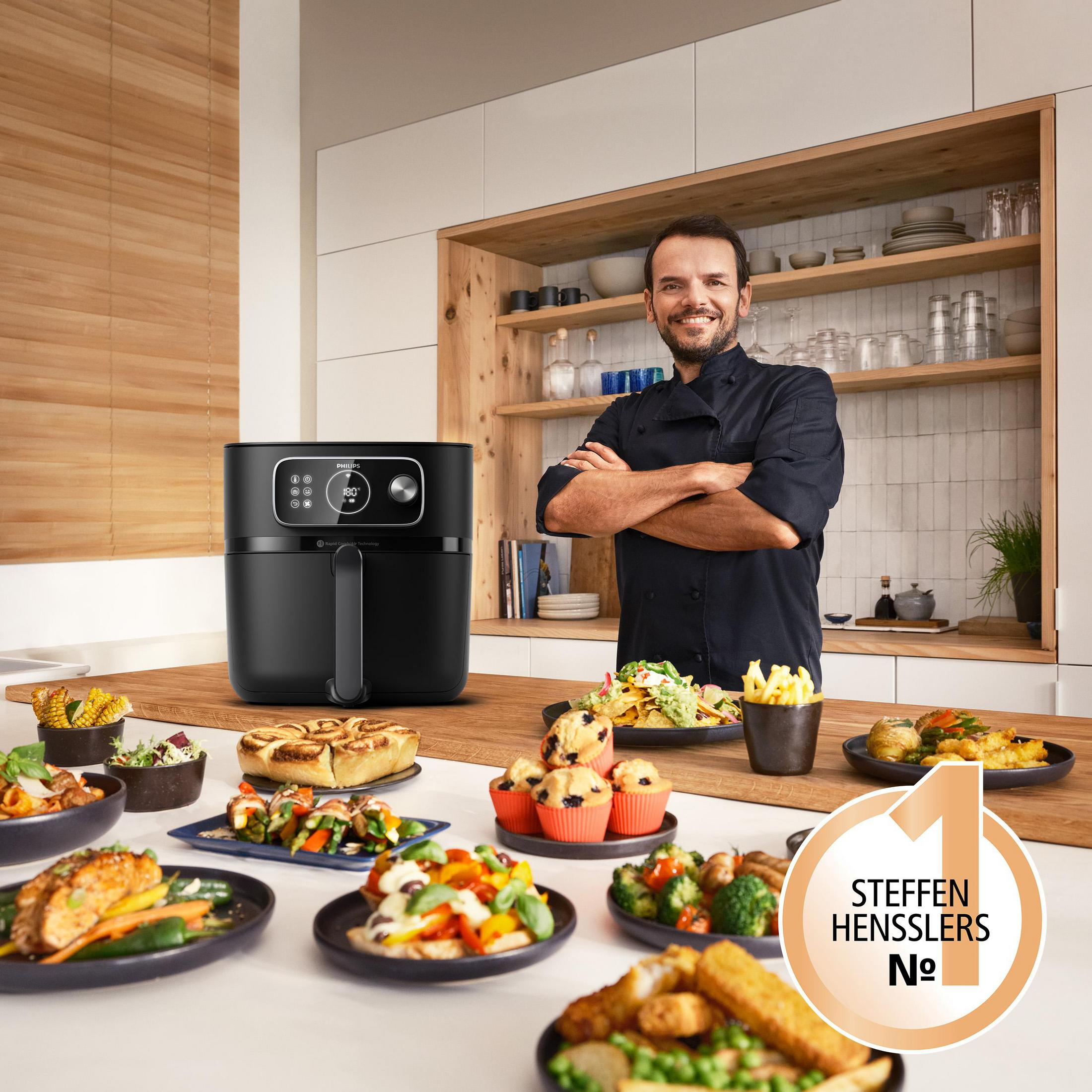 Ein Koch steht mit verschränkten Armen vor einem Tisch voller Speisen und einem schwarzen Airfryer.