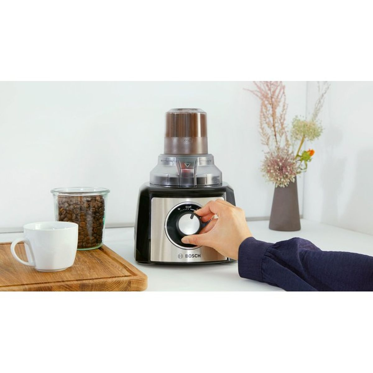 A hand turns the dial on a Bosch grinder next to coffee beans and a mug.