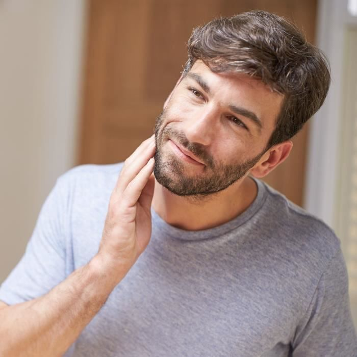 Un hombre con barba se toca la mejilla, viste una camiseta gris.