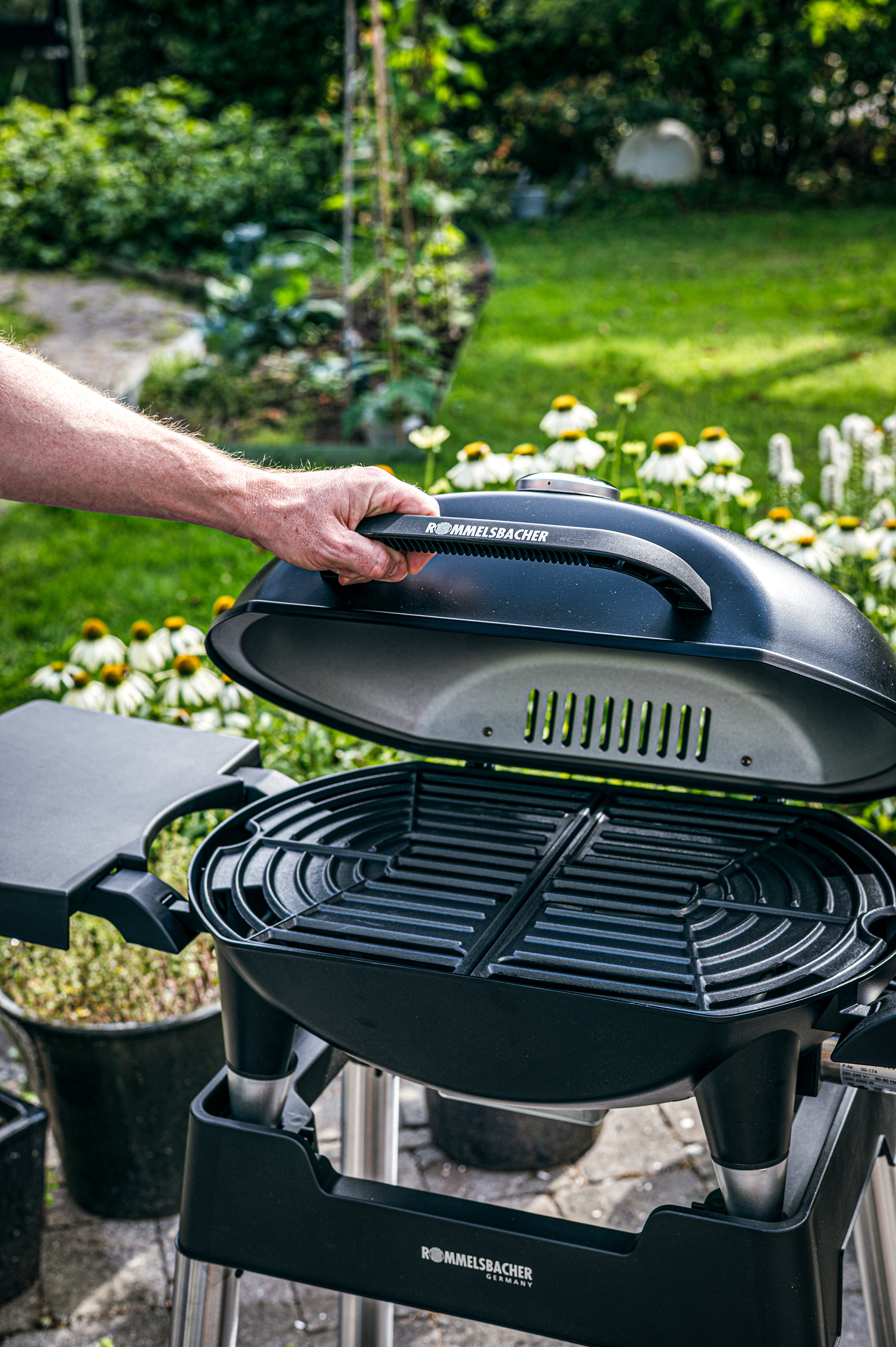 Hand öffnet schwarzen Grill mit Rommelsbacher-Logo im Garten.