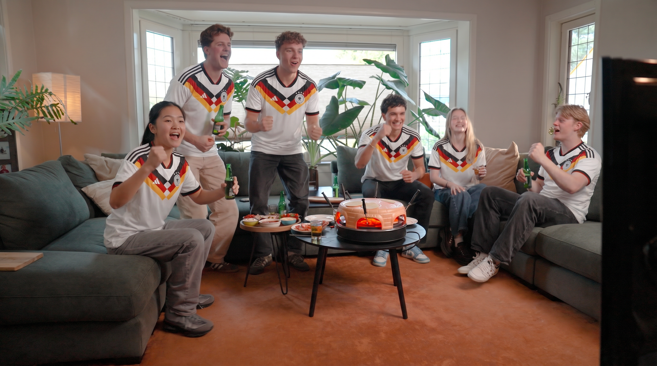 Sechs Personen in Deutschland-Trikots jubeln vor einem Fernseher, mit Snacks.