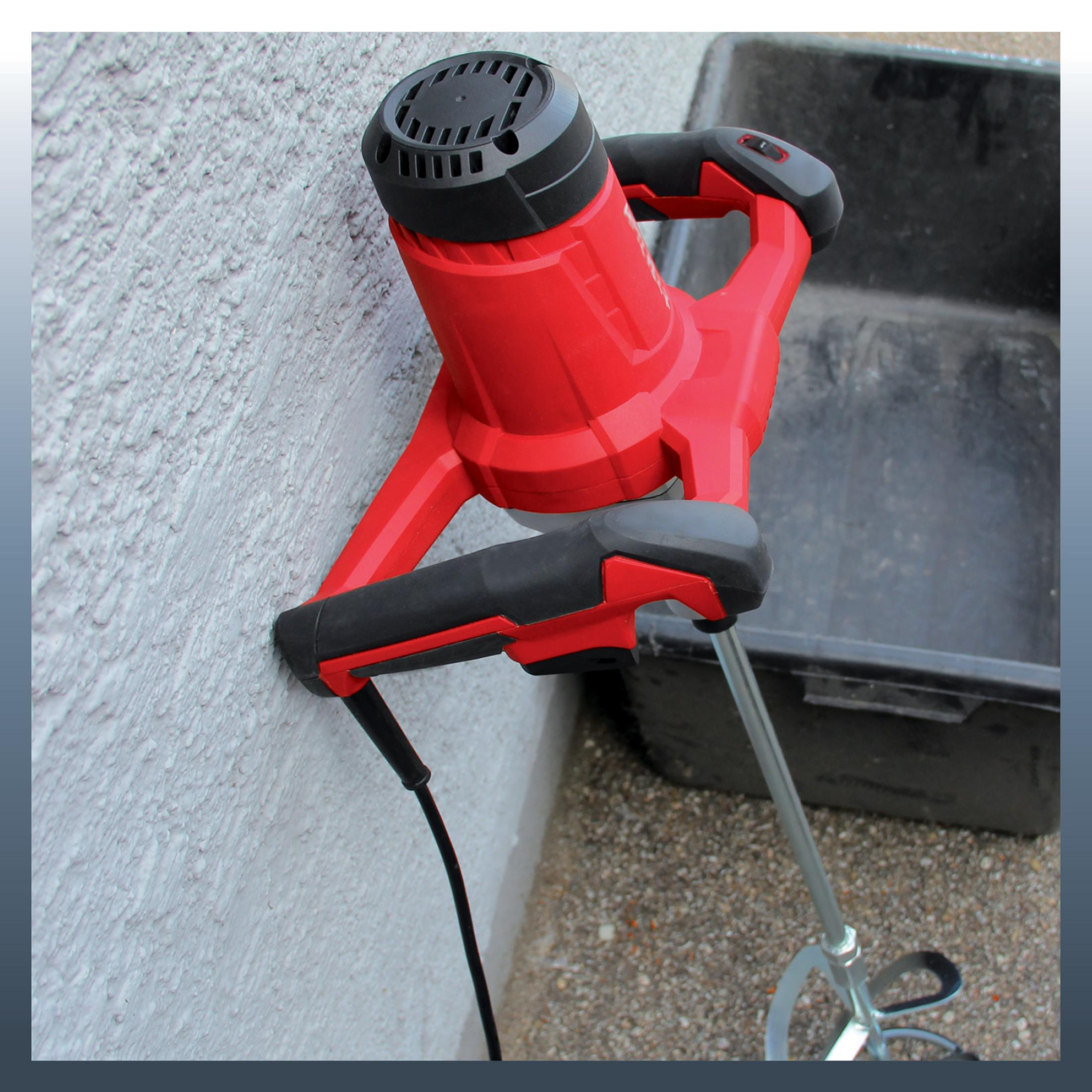 Un taladro mezclador rojo y negro con eje metálico apoyado en pared gris claro rugosa.