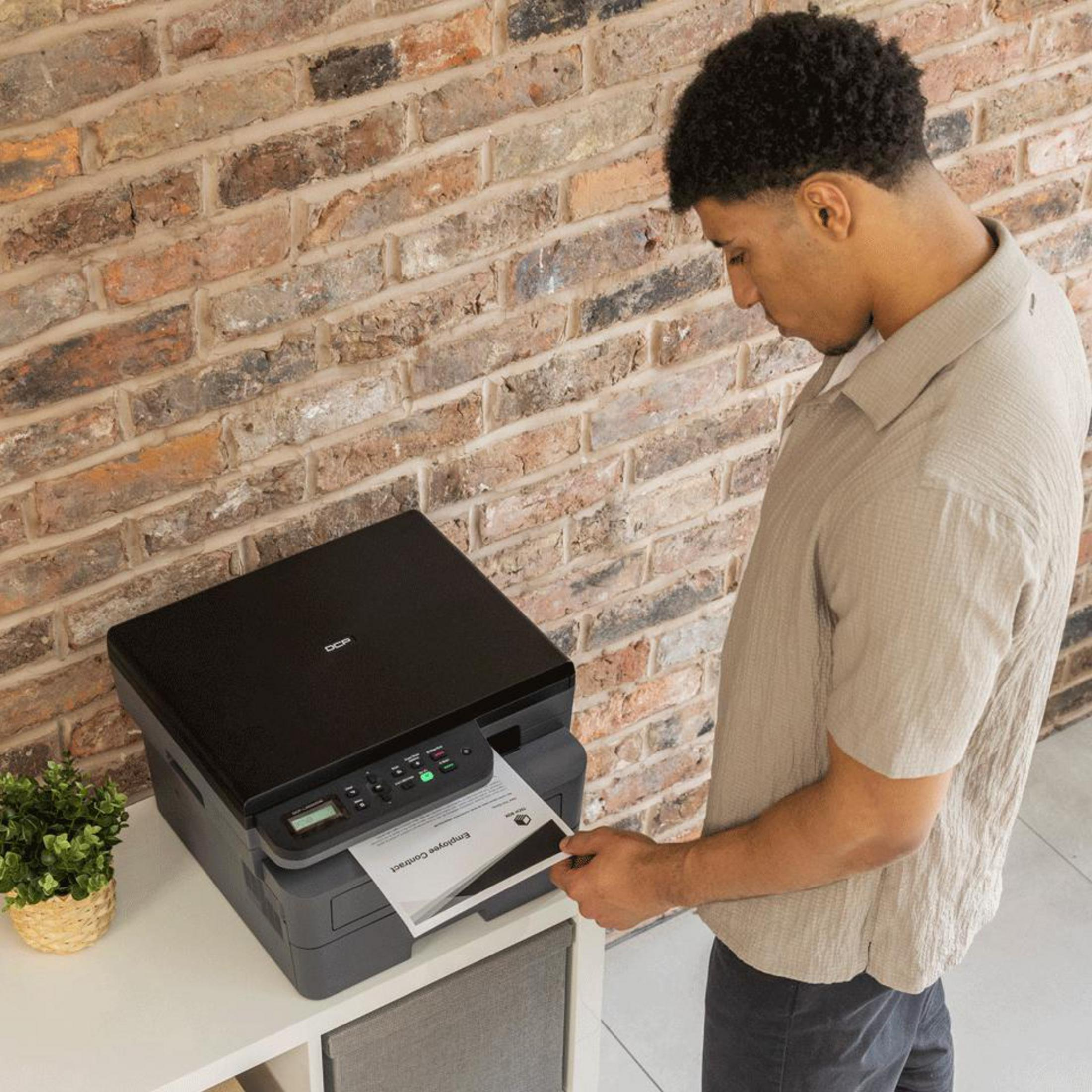 Een man haalt een document uit een printer op een witte kast, met bakstenen muur erachter.