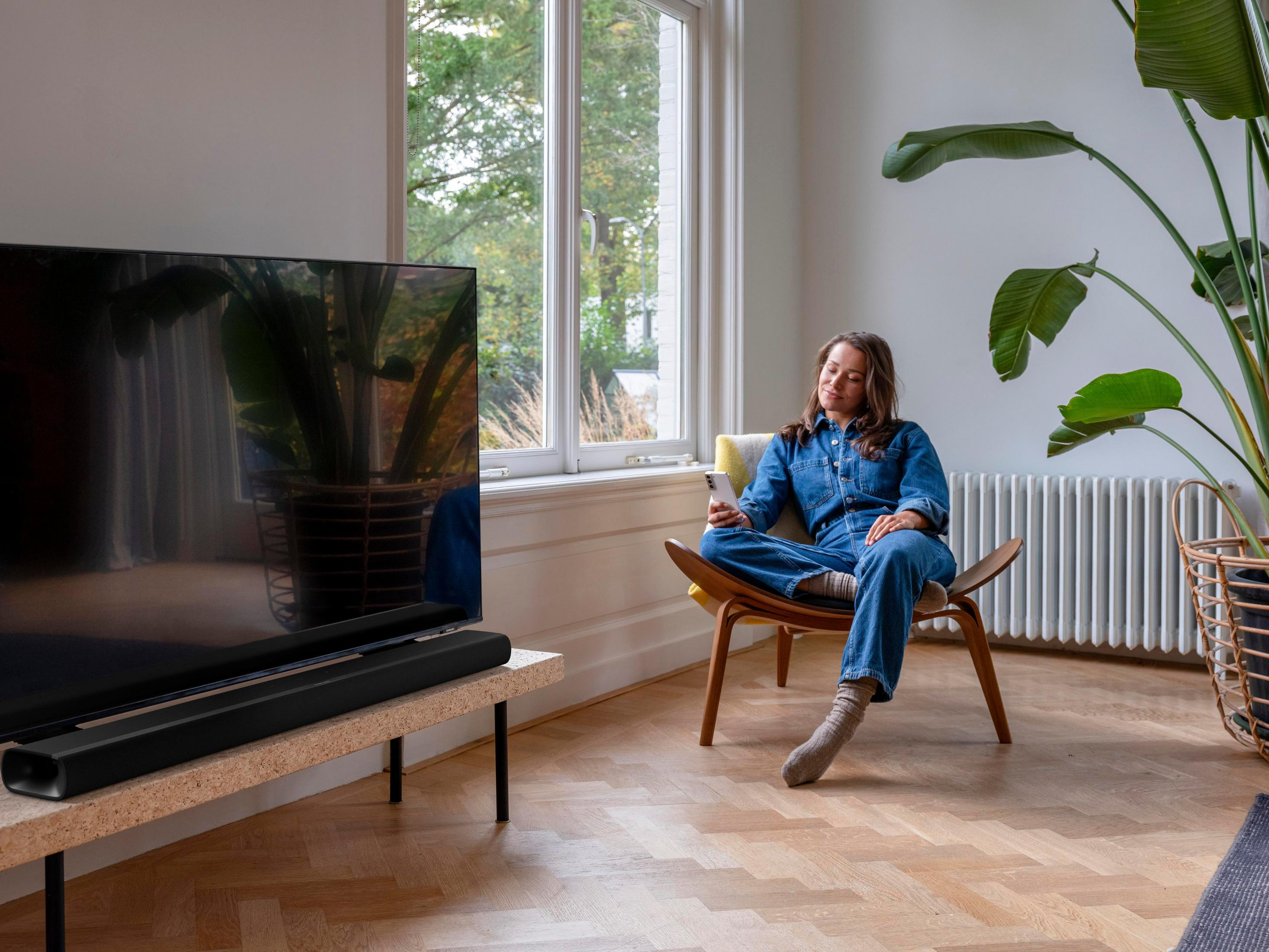 Kobieta na krześle z telefonem przed telewizorem i rośliną.
