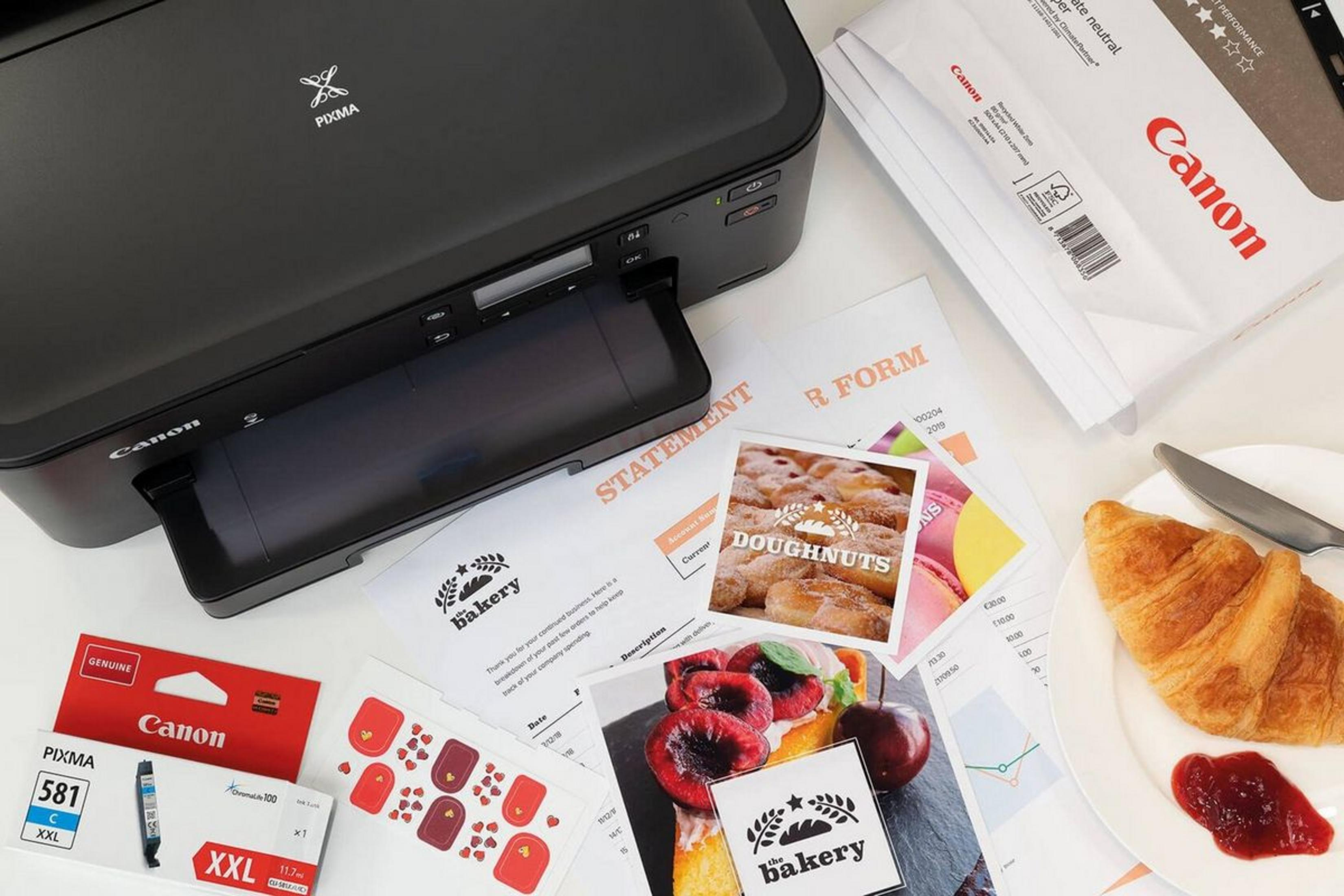 Una impresora Canon Pixma negra con papeles, cartucho de tinta y desayuno.