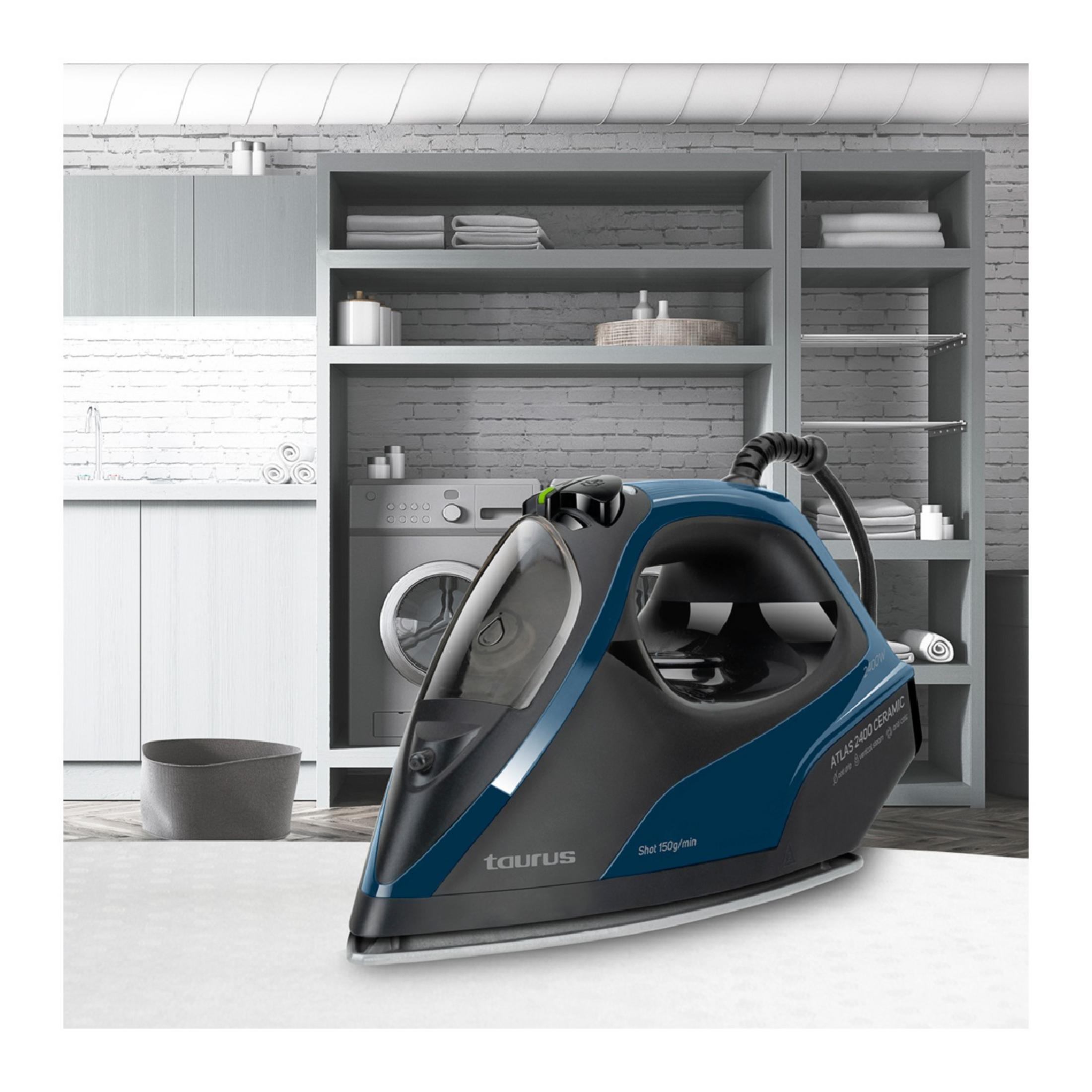 Una plancha de vapor Taurus azul y negra descansa sobre una mesa. Lavadora y estantes al fondo.