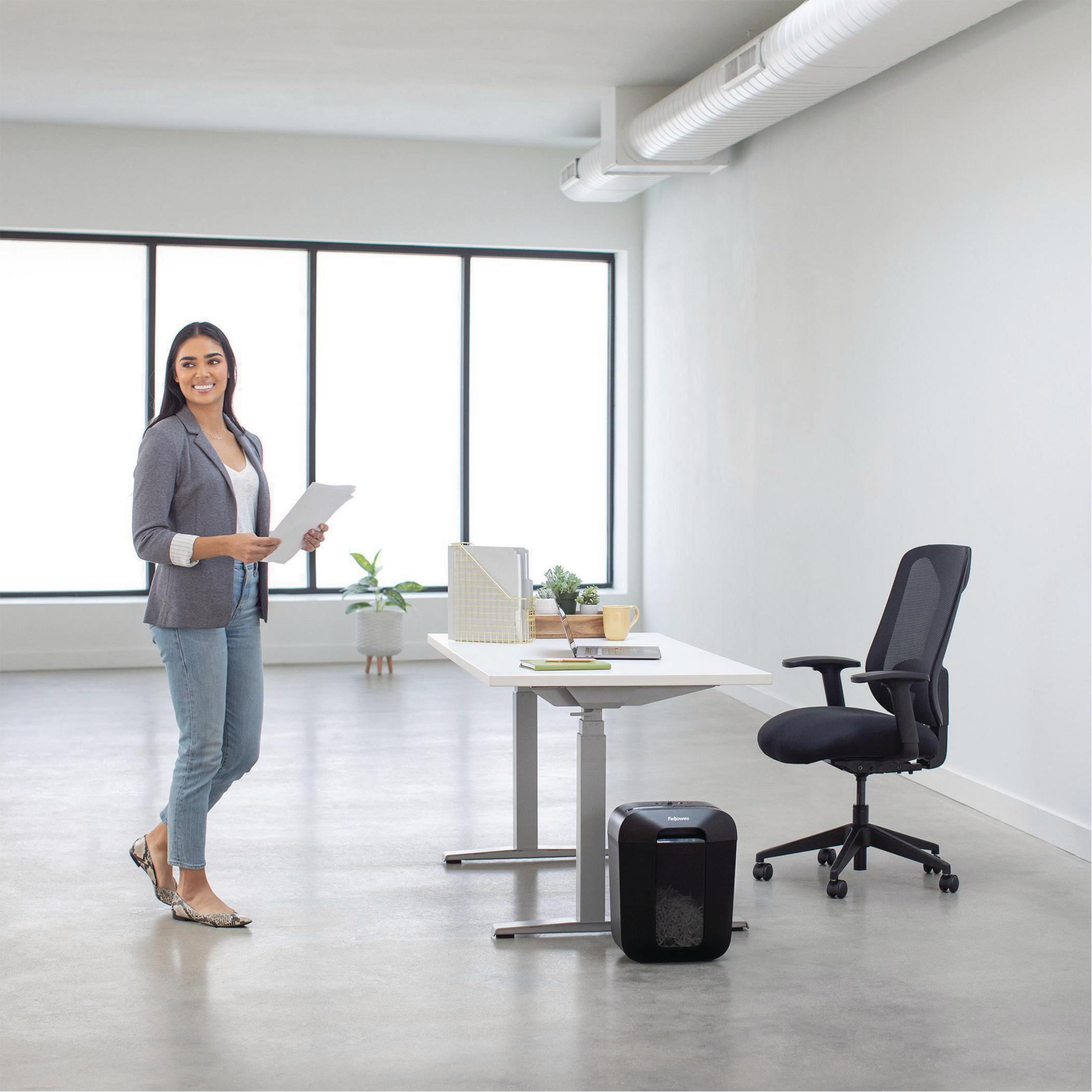 Une femme en blazer tient des papiers dans un bureau avec un bureau et une déchiqueteuse.