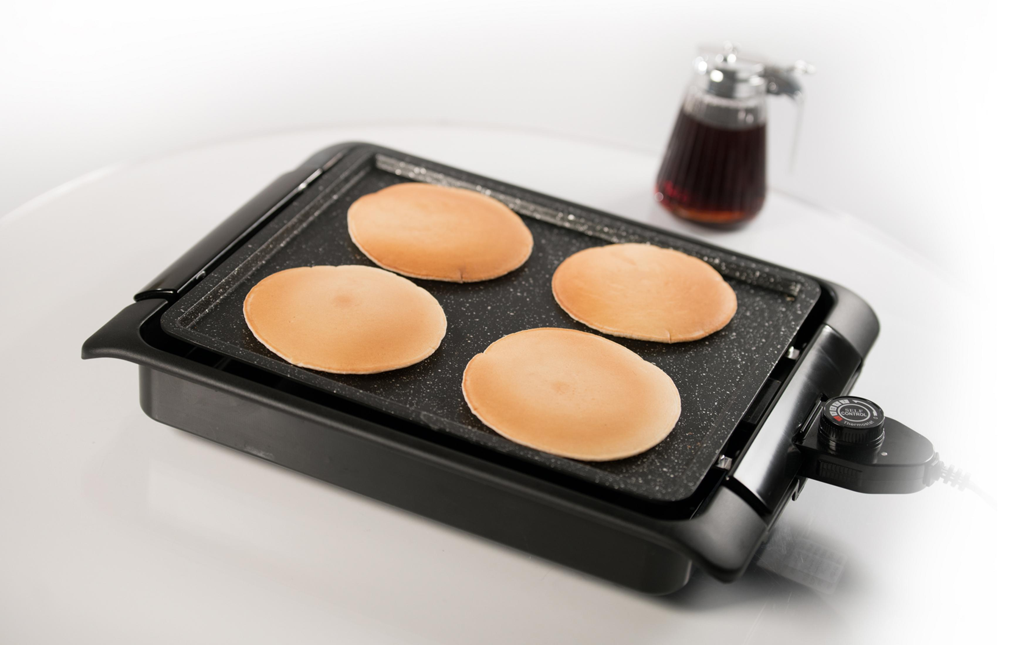 Vier Pfannkuchen auf einer elektrischen Grillplatte neben einem Sirupkrug.