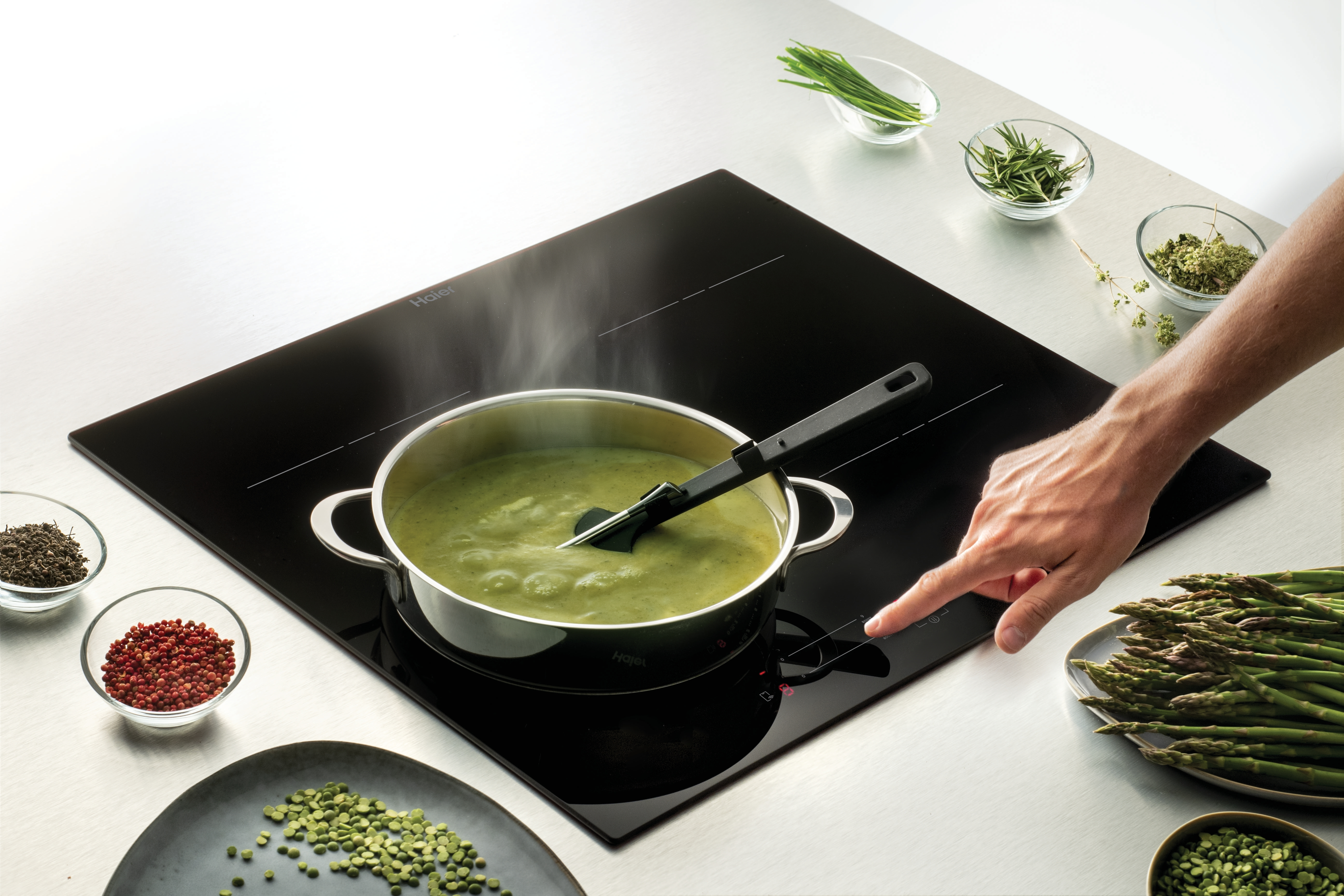 Grüne Suppe kocht in einem Topf auf einem Induktionskochfeld, daneben Spargel und Kräuter.