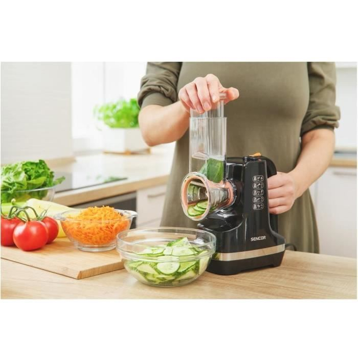 Una persona usa un robot da cucina Sencor nero per affettare cetrioli. Ciotole di cetrioli affettati, carote grattugiate e pomodori sono vicine.