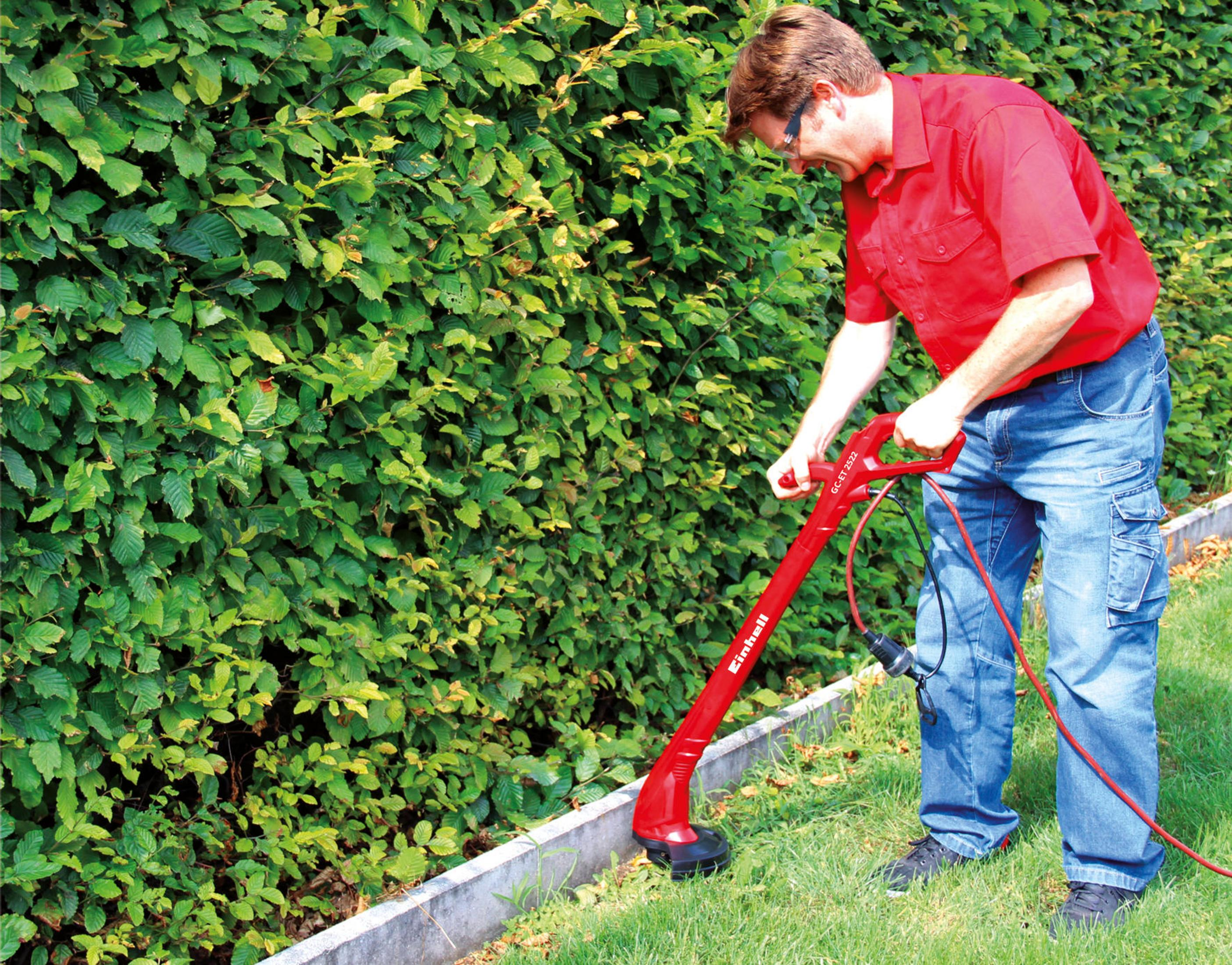 Mann trimmt Hecke mit rotem Rasentrimmer im Garten.