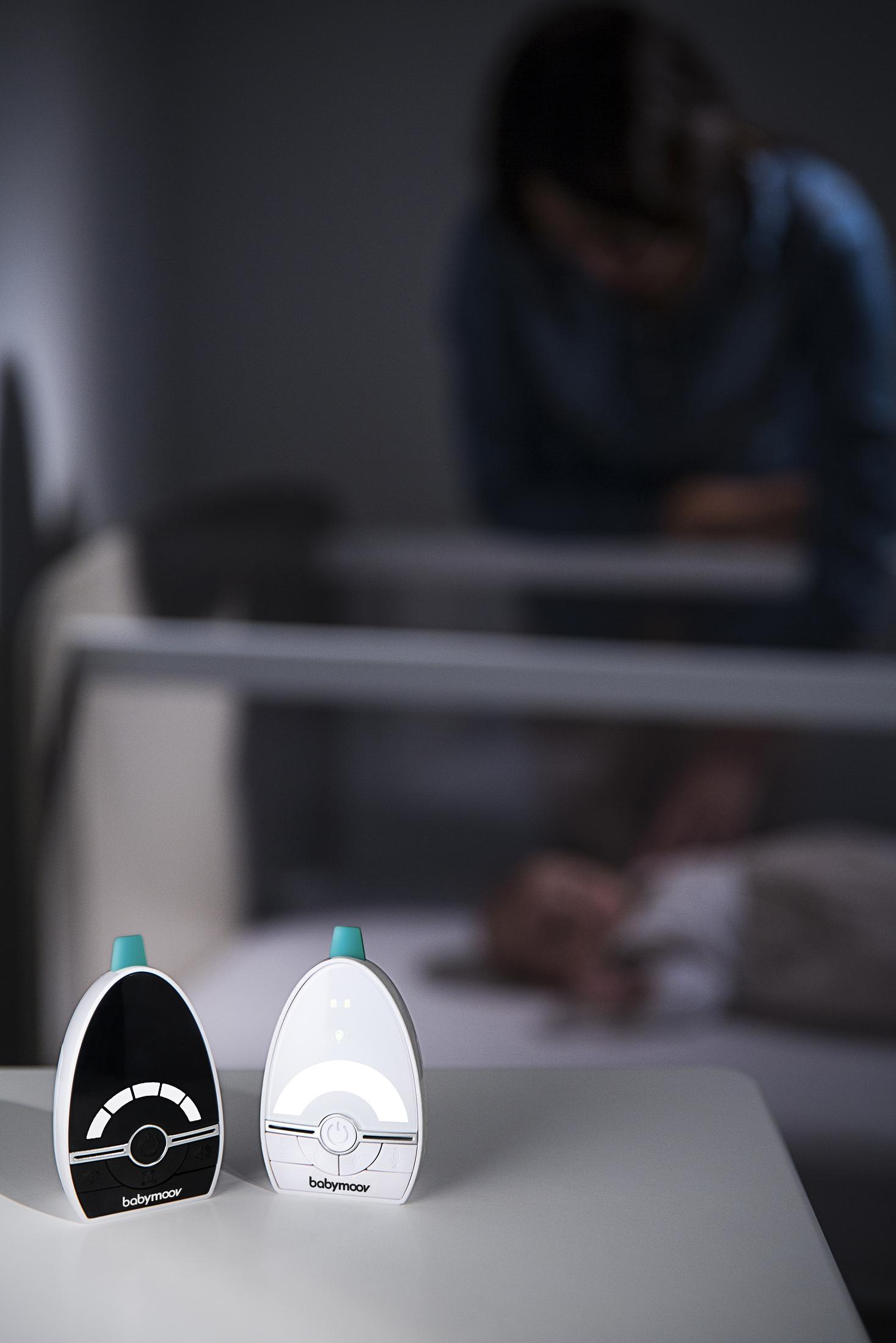 Zwei Babyphones auf einem Tisch vor einem Kinderbett mit einem Baby und einer Person im Hintergrund.