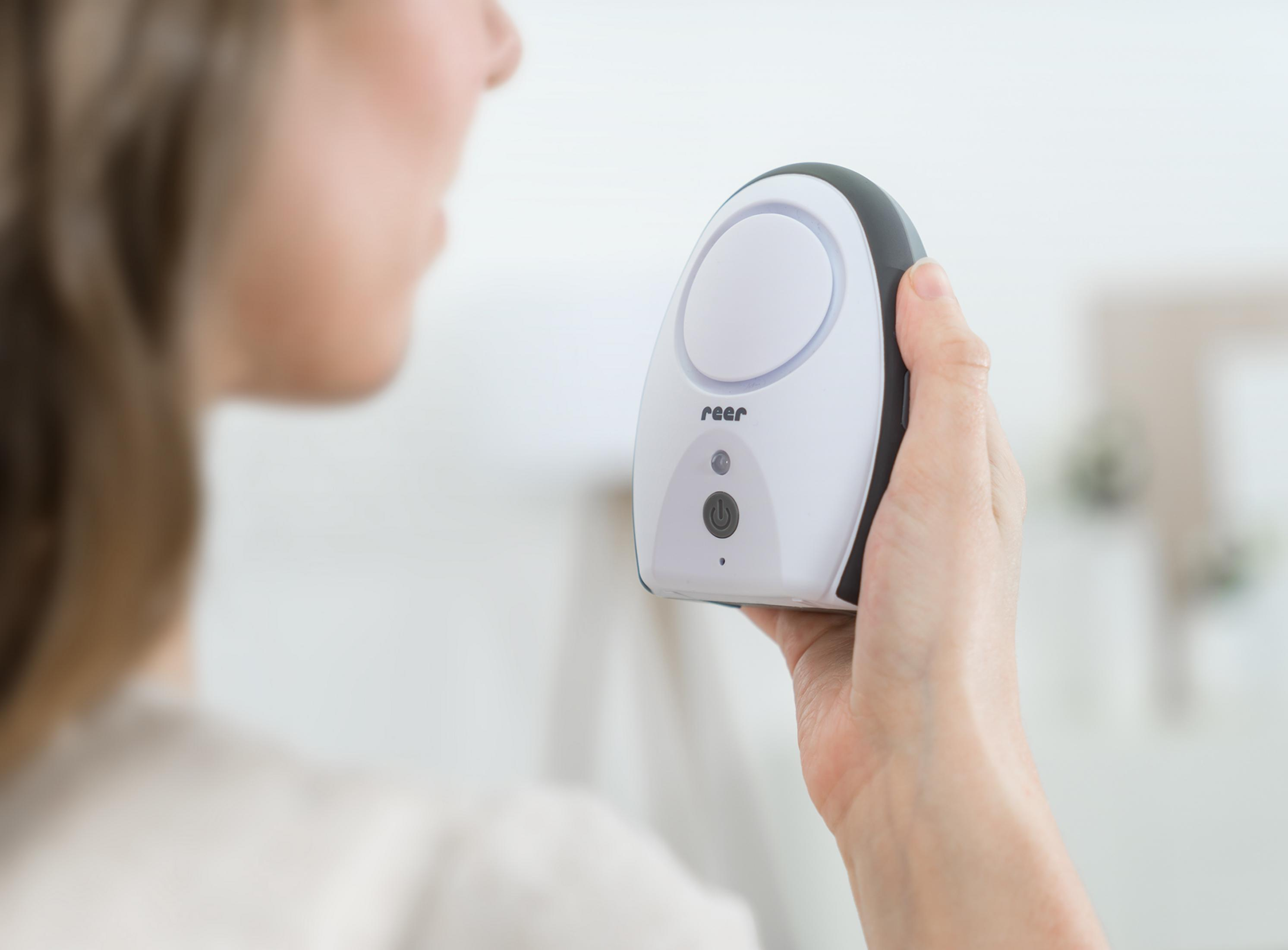 Una donna tiene un baby monitor bianco con accenti grigi. Una luce ad anello è visibile.