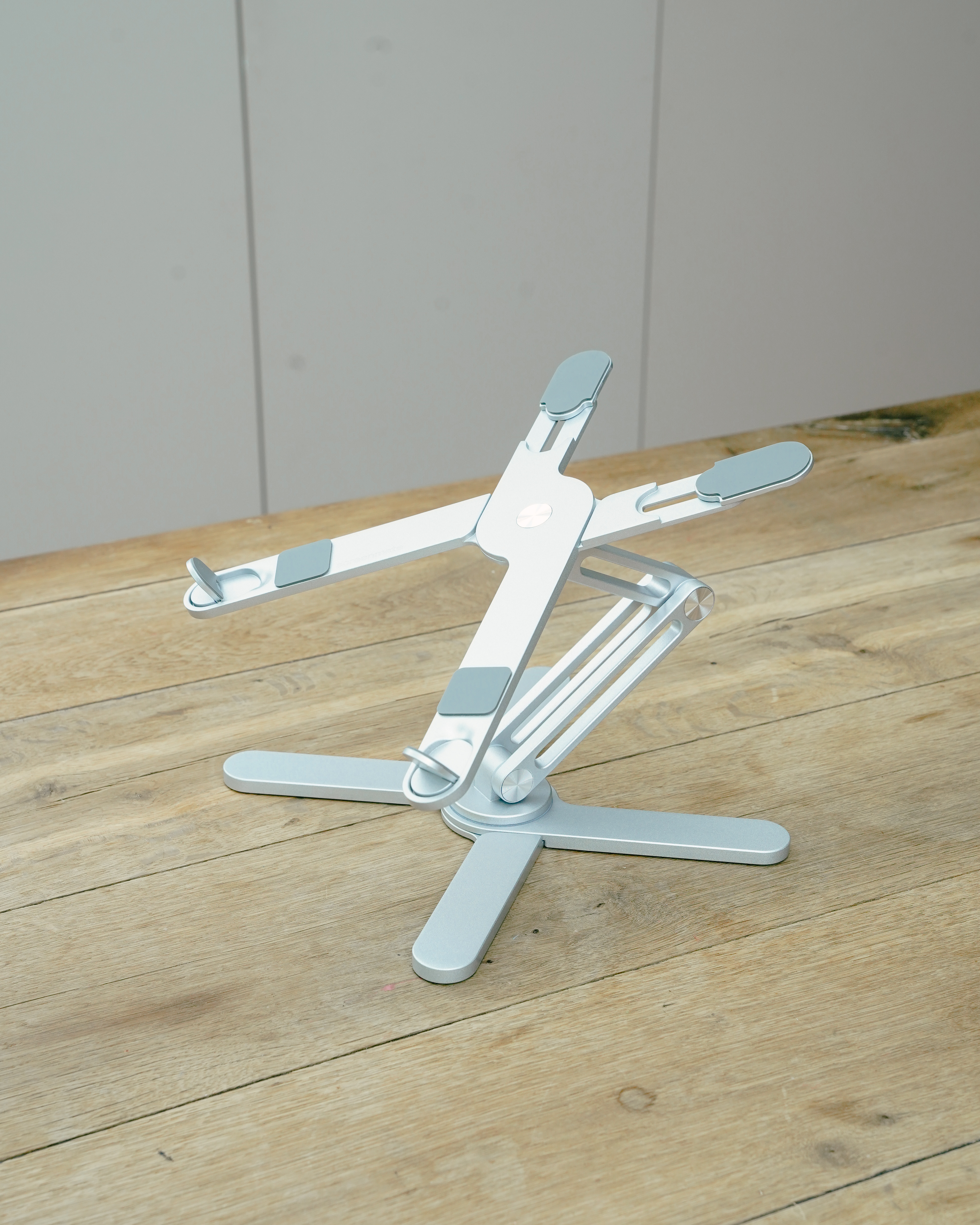 Een zilveren laptopstandaard met verstelbare armen staat op een houten tafel.