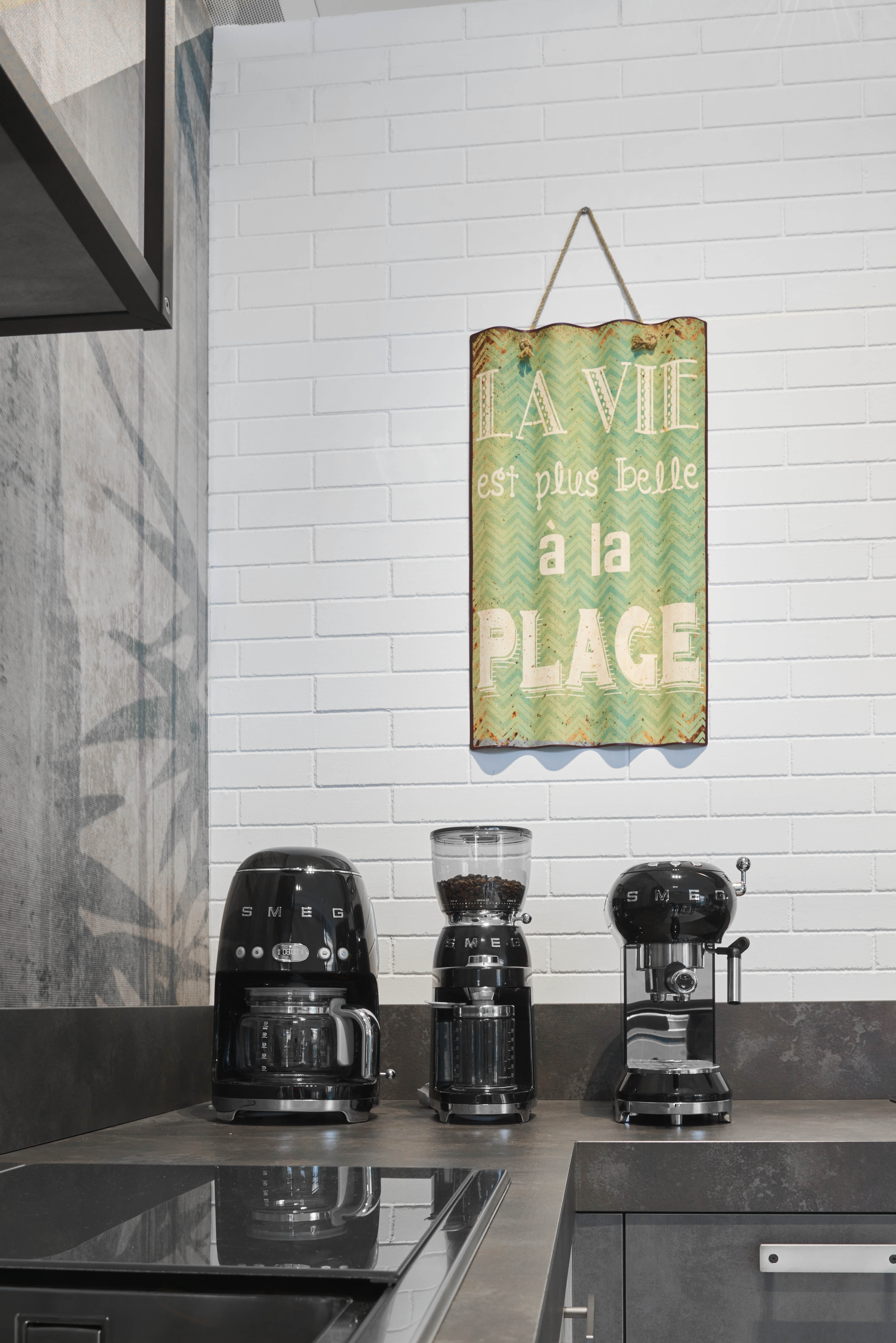 Drei schwarze Smeg-Küchengeräte und ein Schild mit Aufschrift 'La vie est plus belle à la plage'.
