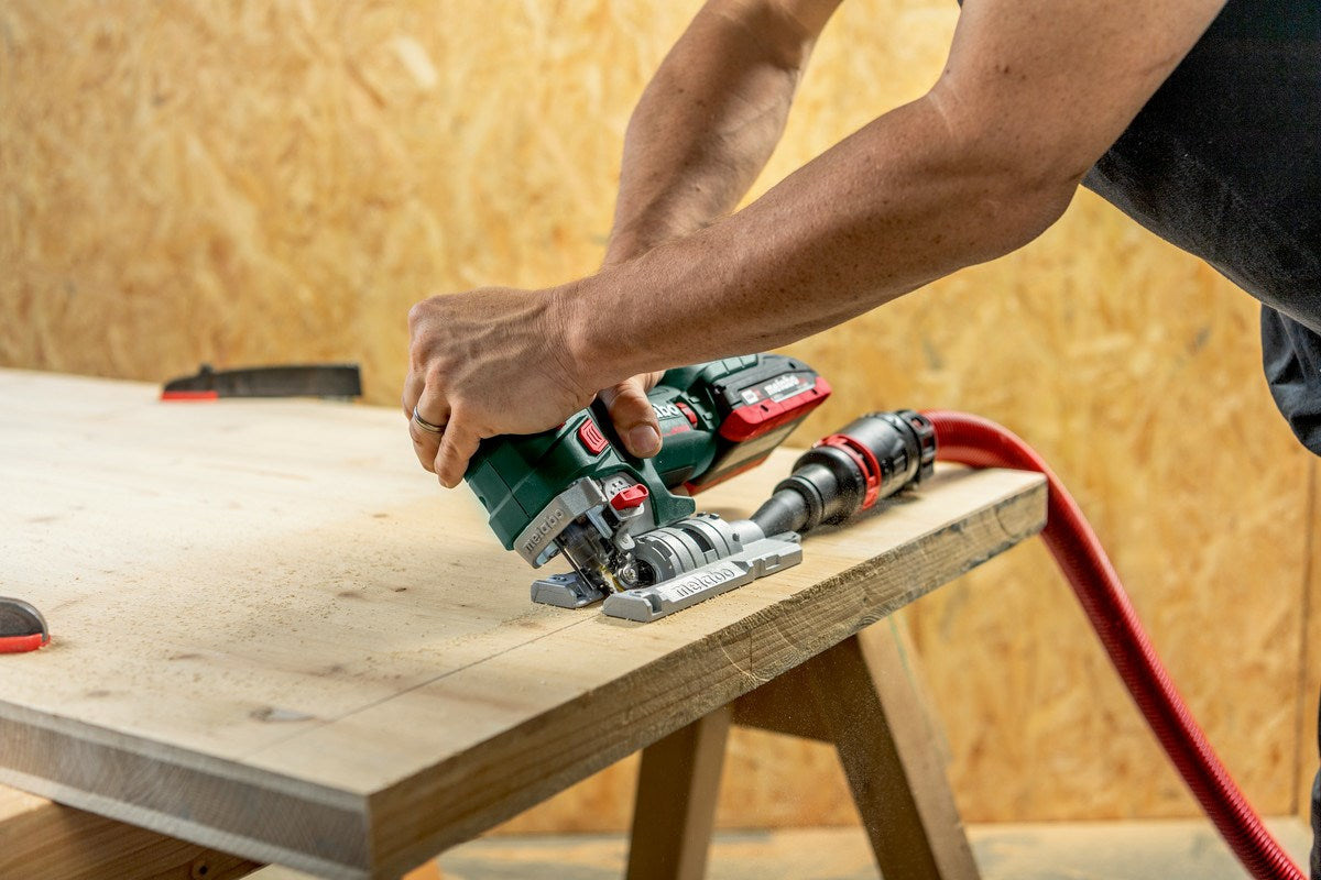 Person cuts wood with a jigsaw, red hose attached.