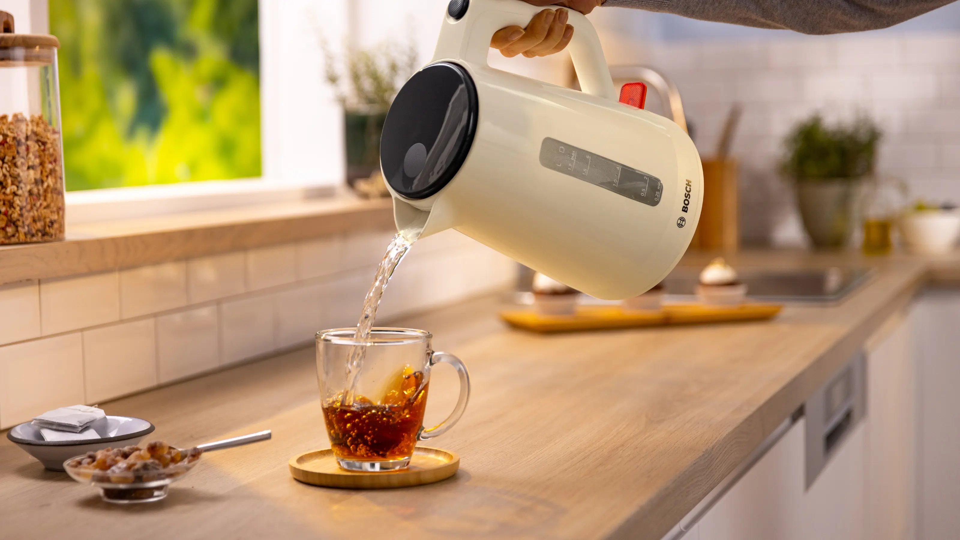 Un bollitore bianco Bosch versa acqua in una tazza di vetro con tè. Cereali, zucchero e bustine di tè sul bancone.