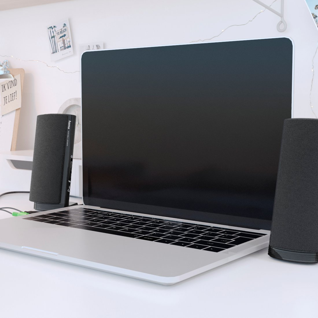 Srebrny laptop z czarnymi głośnikami na białym biurku. Małe notatki i lampki zdobią tło.