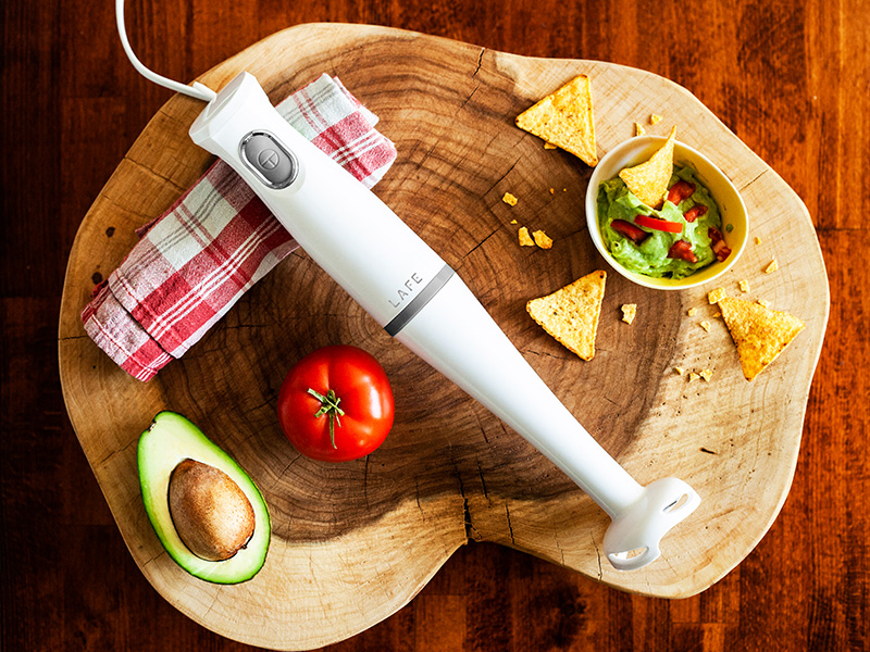 Un frullatore a immersione bianco riposa su un tagliere di legno vicino ad avocado, pomodoro, guacamole e patatine.