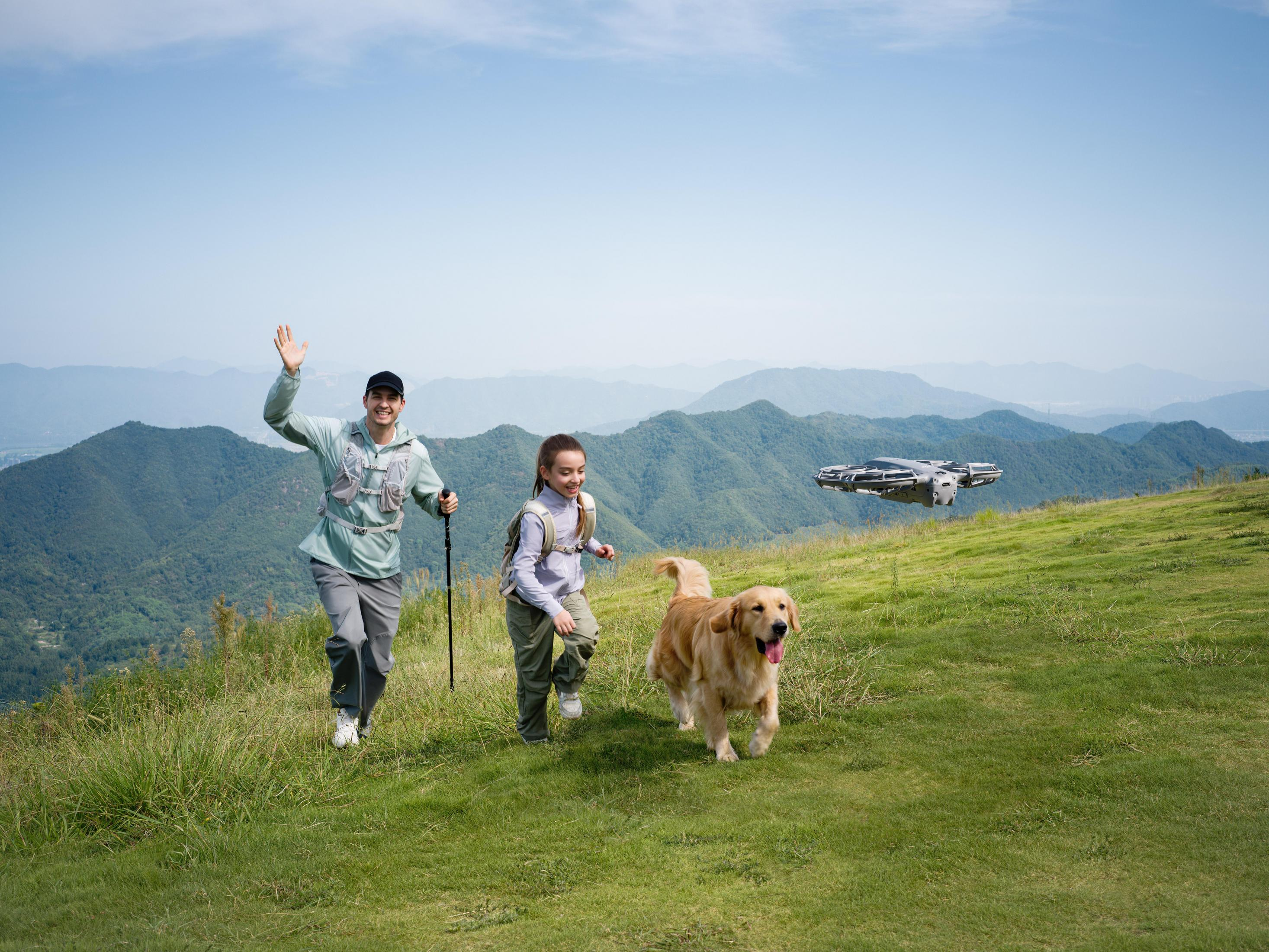 Hombre saluda, corre con niña y perro en colina con dron y montañas.