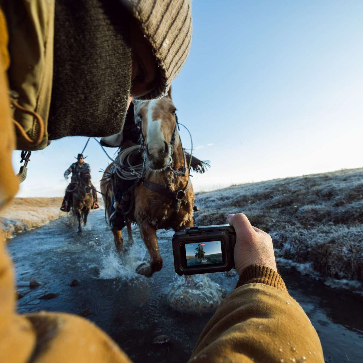 Un hombre filma a dos vaqueros cabalgando a través de un arroyo.