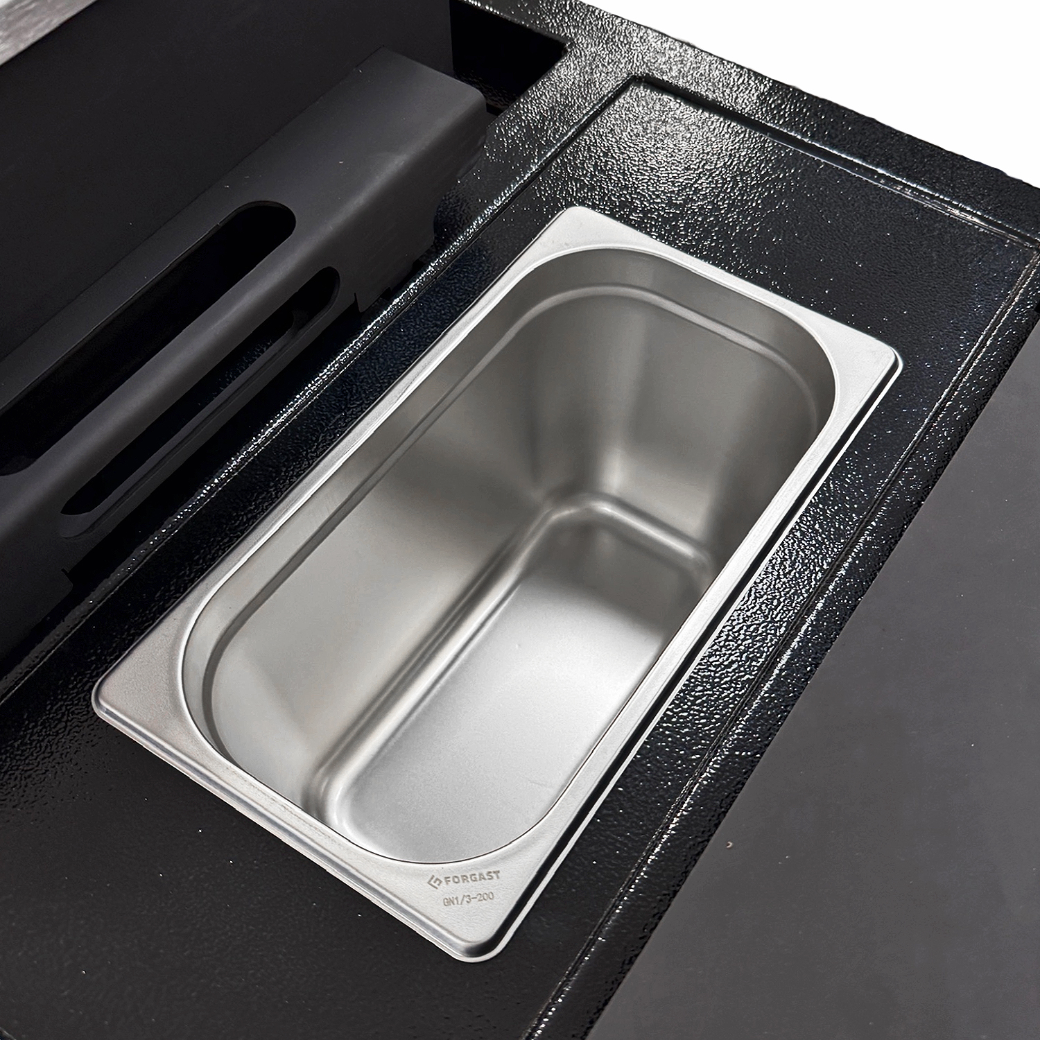 Eine Metall-Chafing Dishschale sitzt in einer schwarzen, texturierten Vertiefung. Ein schwarzer Kunststoffgriff ist sichtbar.