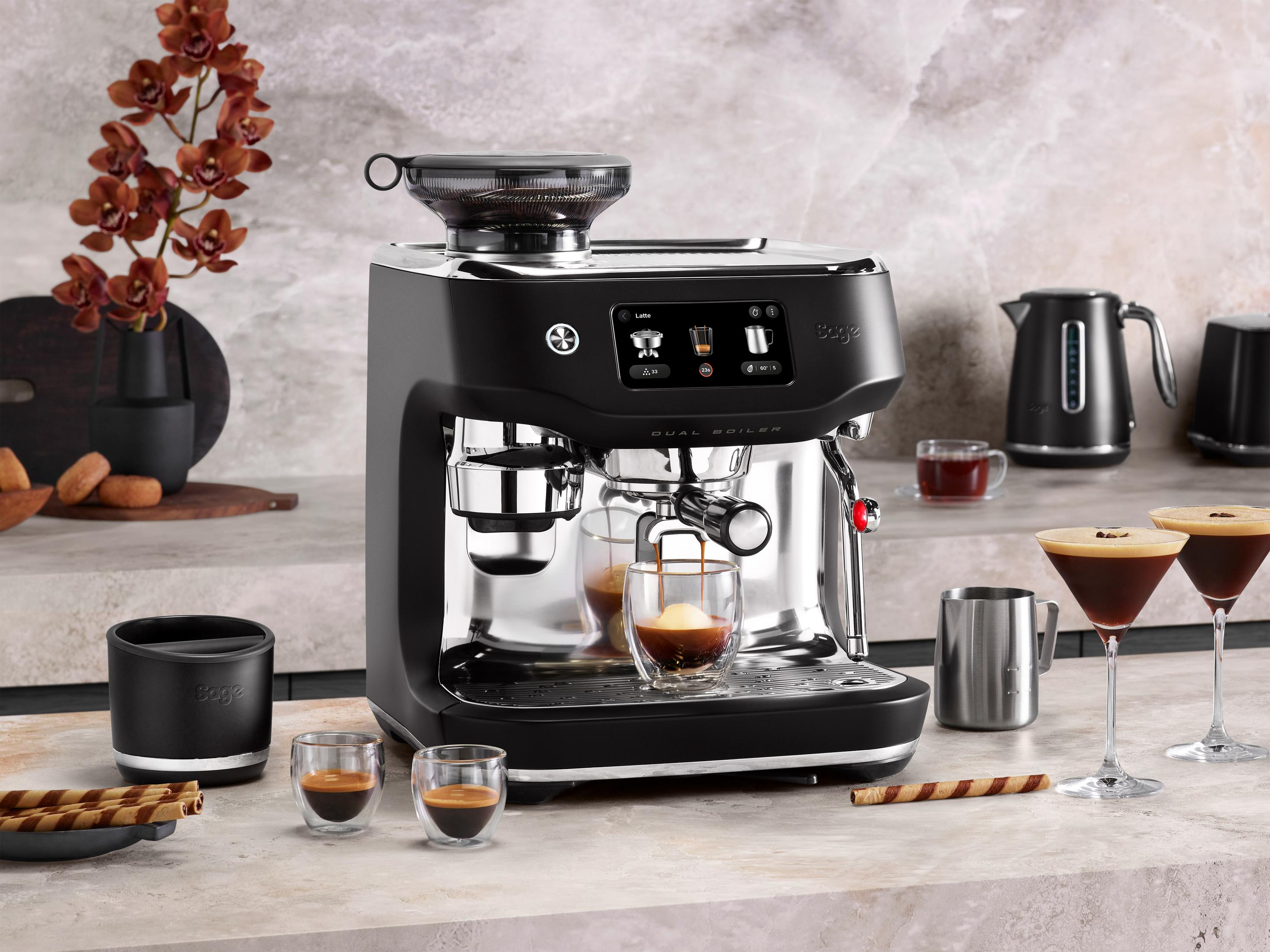 Una macchina per espresso nera con montalatte, tazzine da espresso, biscotti e fiori.