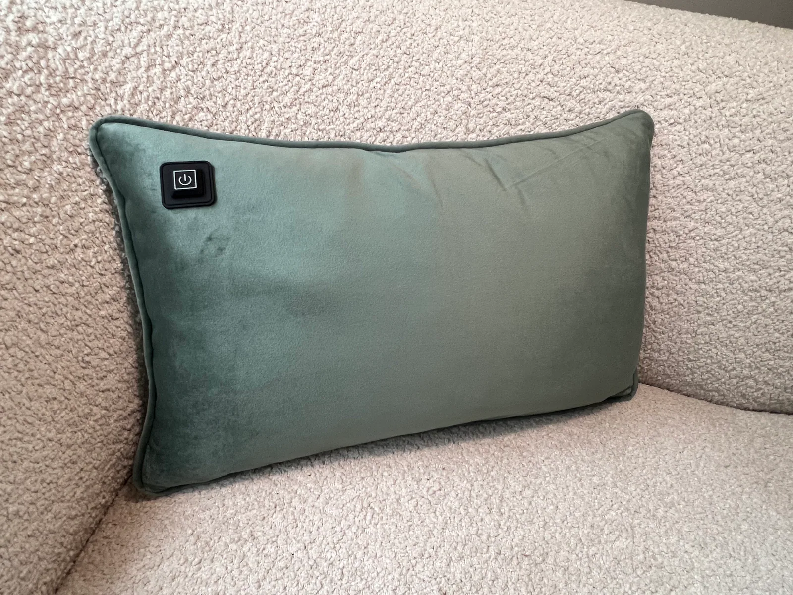 Ein grünes Kissen mit einem schwarzen Ein-/Ausschaltknopf liegt auf einem strukturierten hellen Sofa.